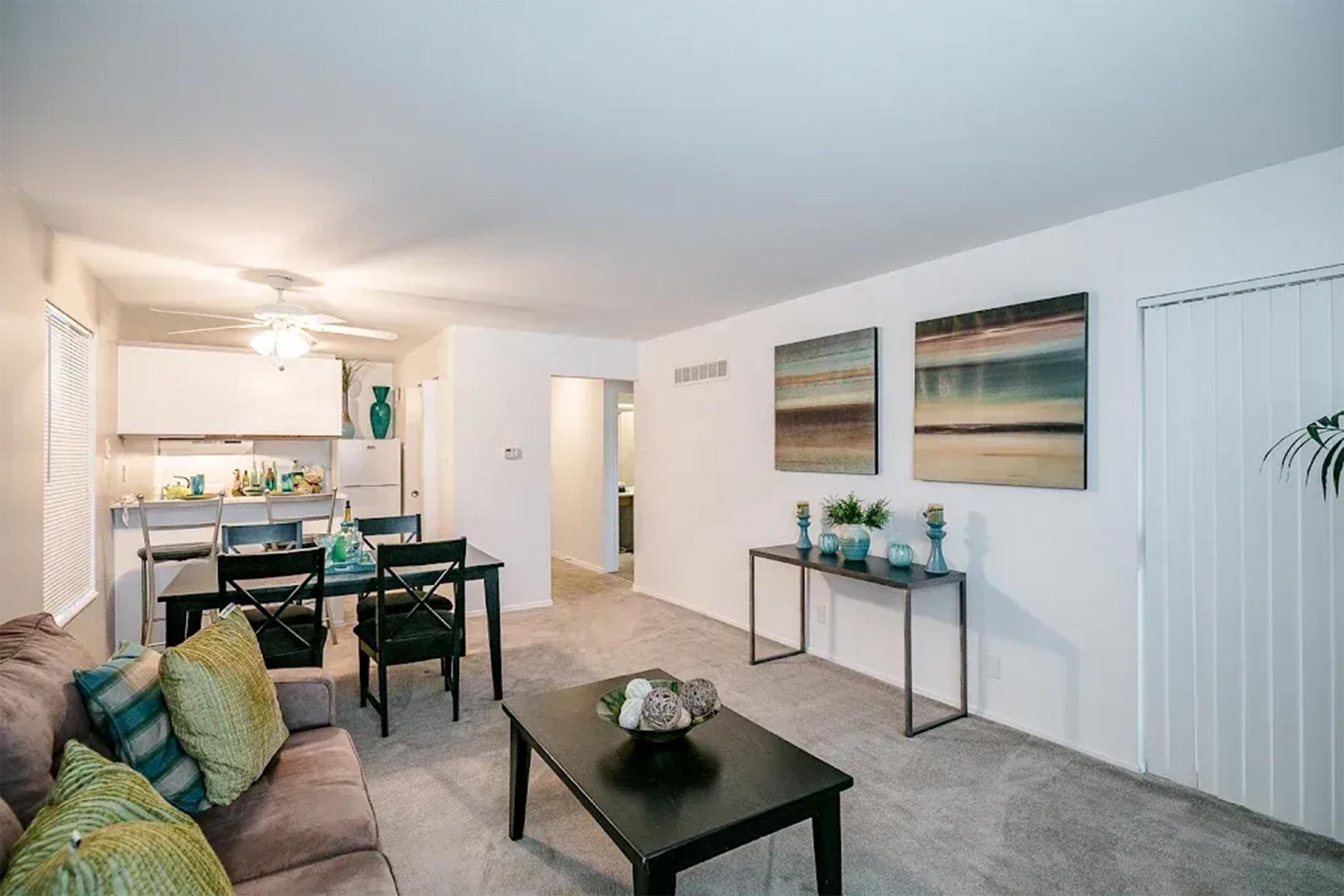A cozy living room featuring a brown couch with green and yellow pillows, a black coffee table with decorative items, and a dining area with a black table and chairs. The walls are adorned with two large abstract paintings, and there's a small kitchen visible in the background. The space is well-lit and inviting.