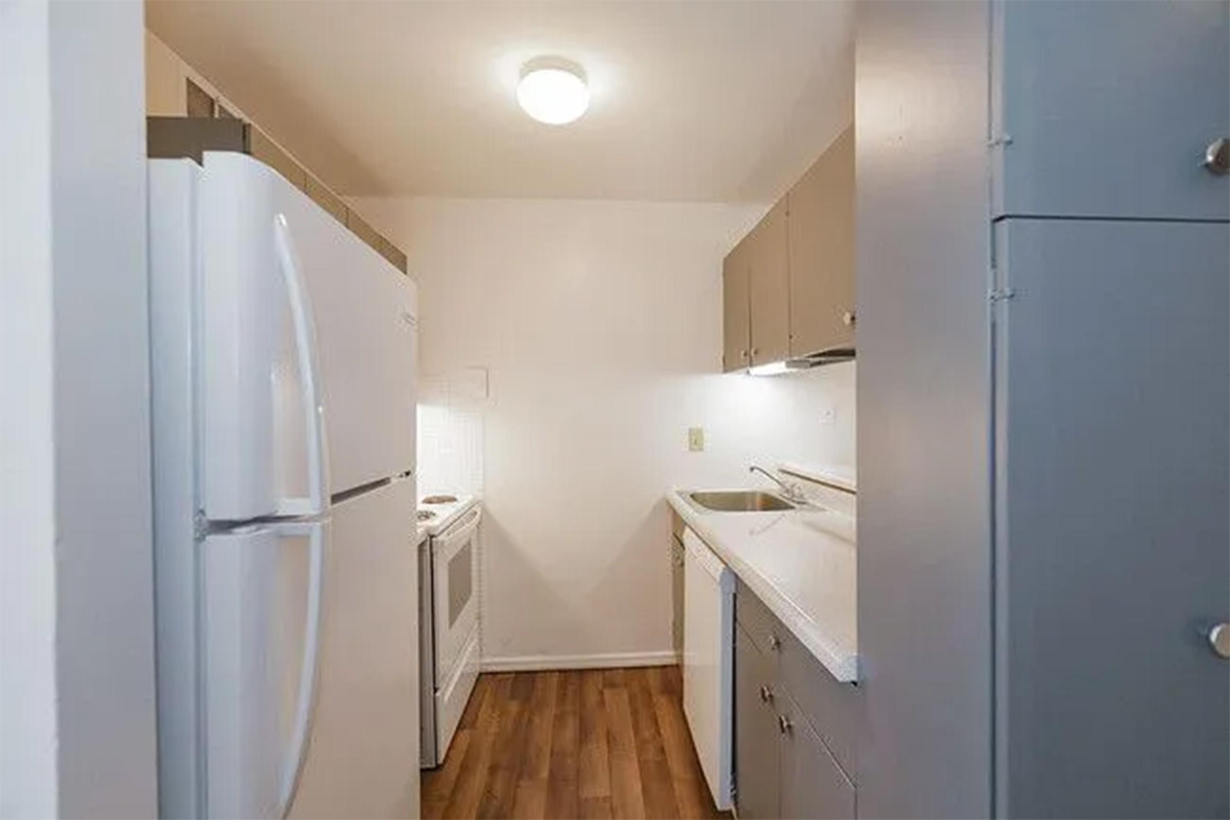 A small, modern kitchen featuring white appliances including a refrigerator and stove. The cabinetry is light gray, with a sink and countertop visible. The floor is made of wood laminate, and the room is well-lit by overhead lighting. The walls are painted a light color, creating a clean and inviting atmosphere.