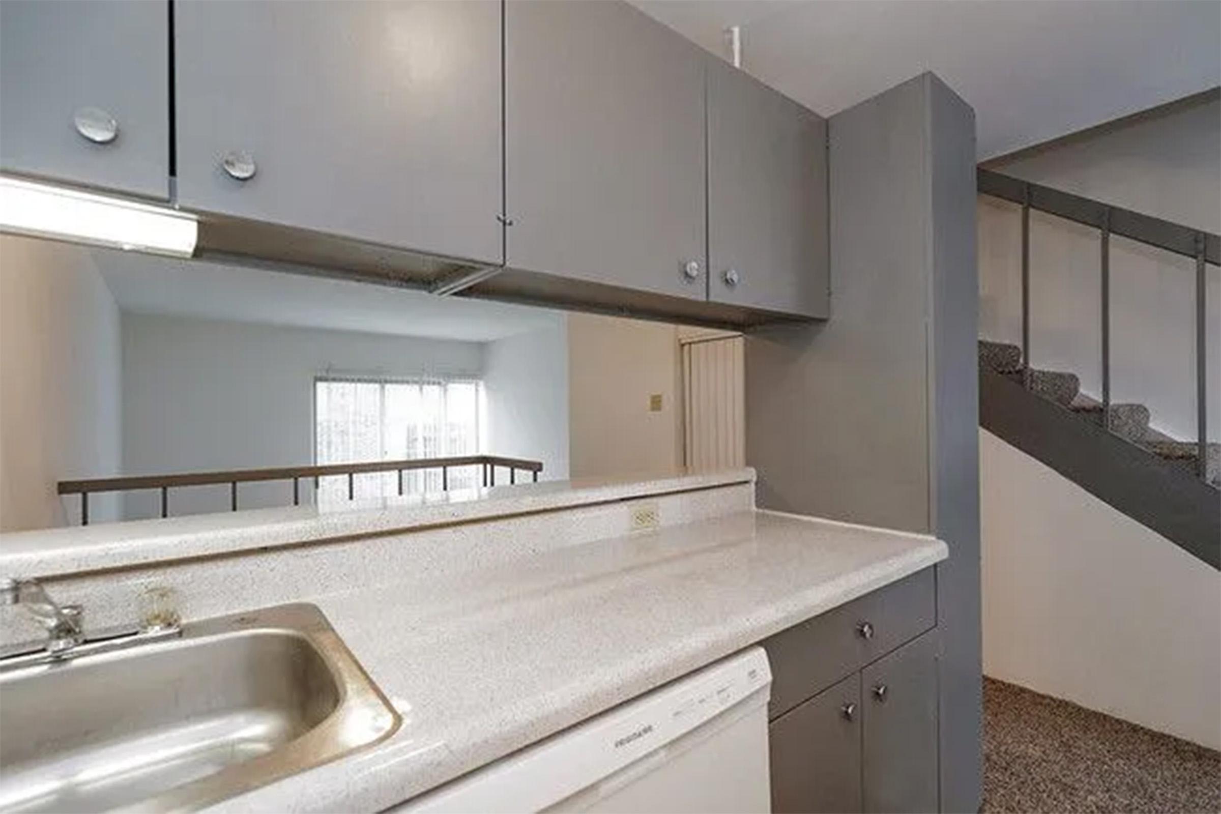 A modern kitchen area featuring a countertop with a sink, a dishwasher, and overhead cabinets. In the background, there is a staircase leading to an upper level, and large windows providing natural light into the space. The flooring is carpeted, contributing to a cozy ambiance.