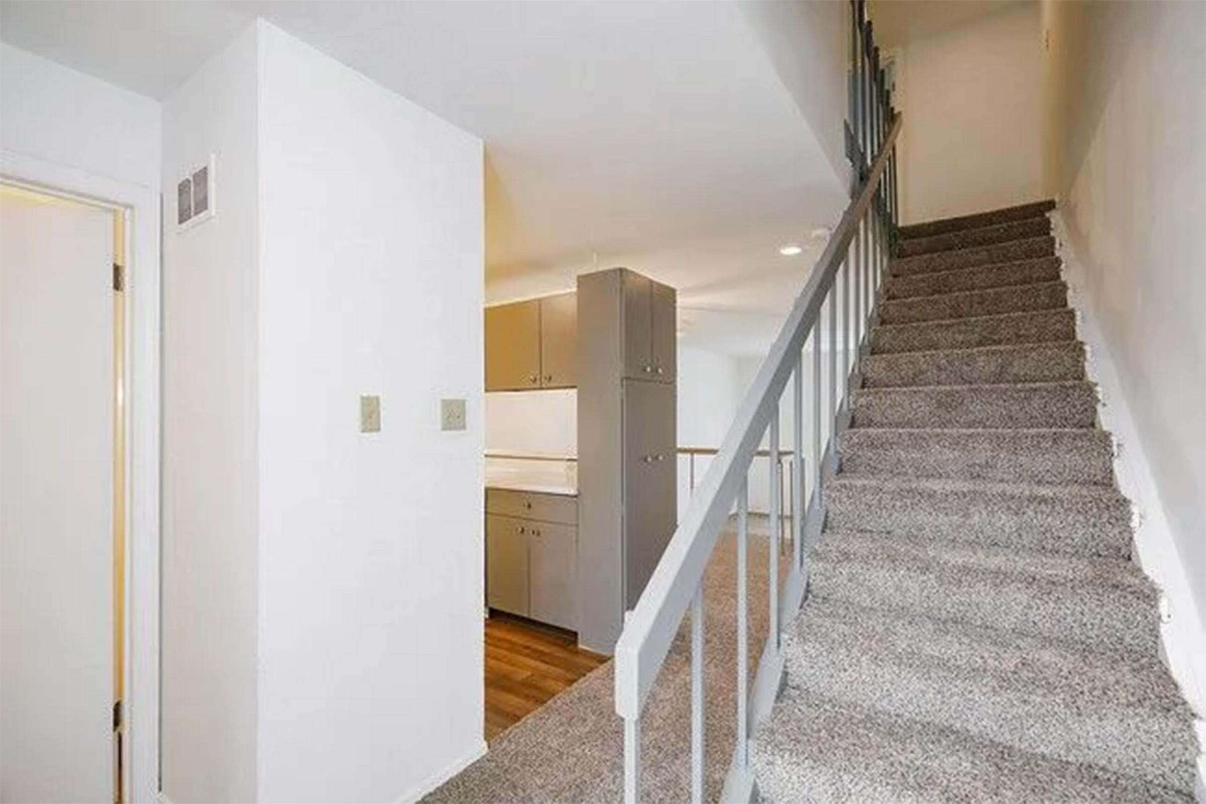 A stairway leading to an upper level, with carpeted steps and a light-colored wall. At the top of the stairs, a kitchen area with cabinets can be seen. The space is bright and modern, featuring an open design that connects the staircase to the kitchen.