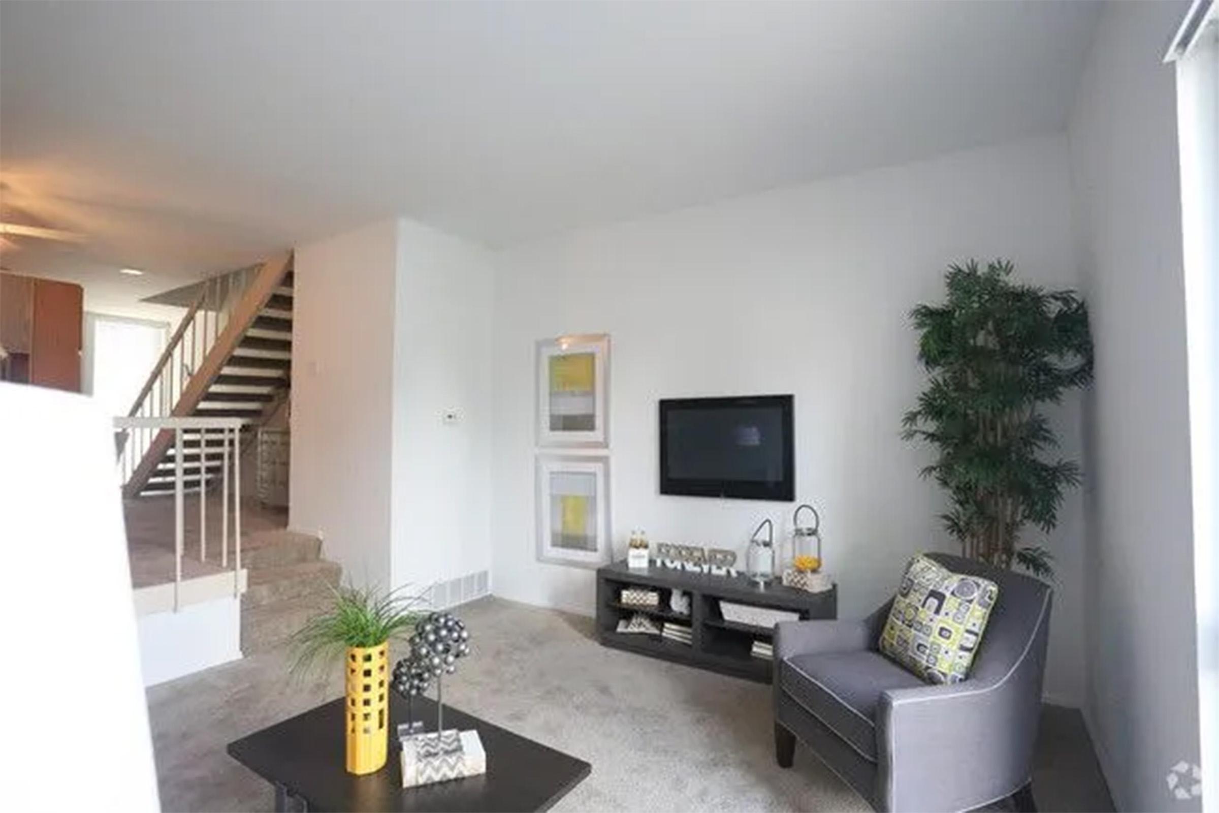 A modern living room featuring a gray armchair, a coffee table with decorative objects, and a television mounted on the wall. The room has a potted plant, two framed artworks, and a staircase in the background. The walls are painted white, and the flooring appears to be carpeted.