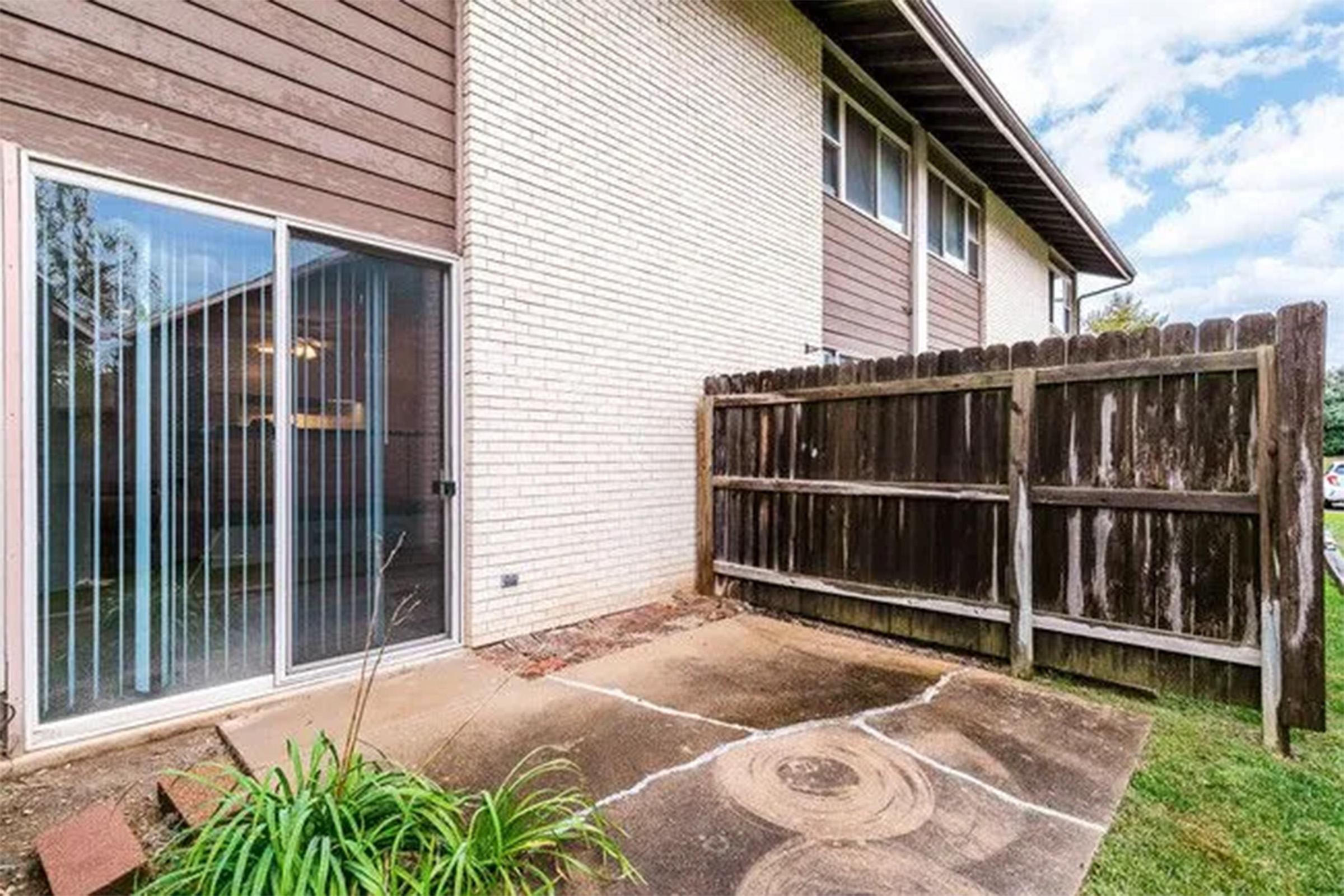 A patio area with a sliding glass door leading inside. The flooring is concrete with visible cracks, surrounded by sparse green plants. A wooden fence borders the space, which appears to be part of a residential building with a two-tone exterior. The sky is partly cloudy.