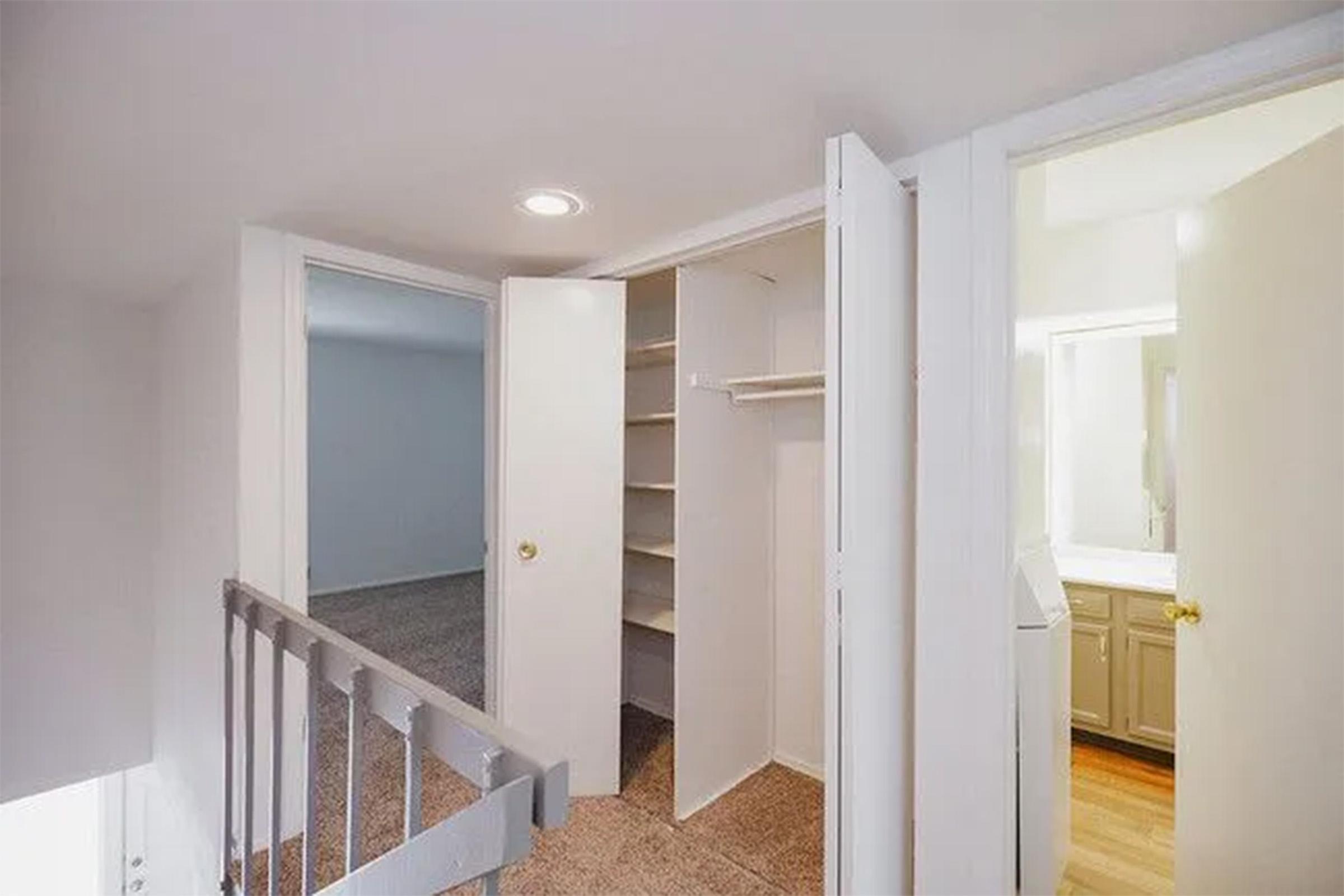 A small hallway leading to two open doors. The left door reveals a closet with shelves, while the right door opens into a bathroom with a vanity. The walls are painted white, and the flooring appears to be light-colored carpet. Natural light is provided by a ceiling fixture.