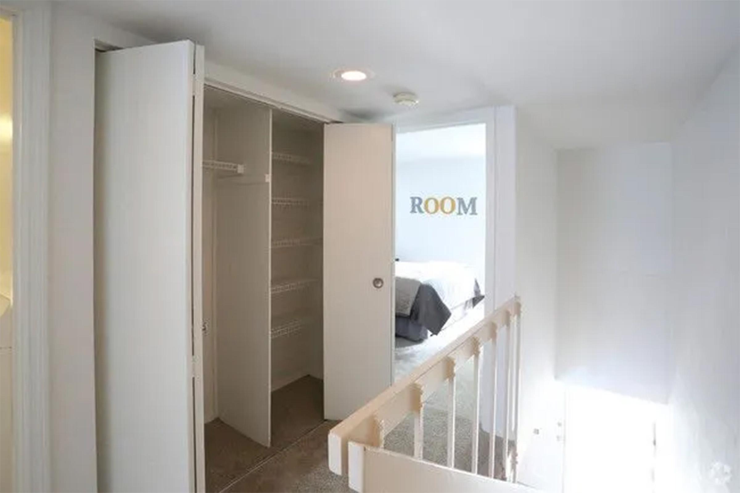 A bright, minimalist hallway leading to a room, with two open white closet doors revealing shelves inside. The wall on the side has the word "ROOM" in gold lettering. Light enters from the left, creating a welcoming atmosphere in a modern interior space.