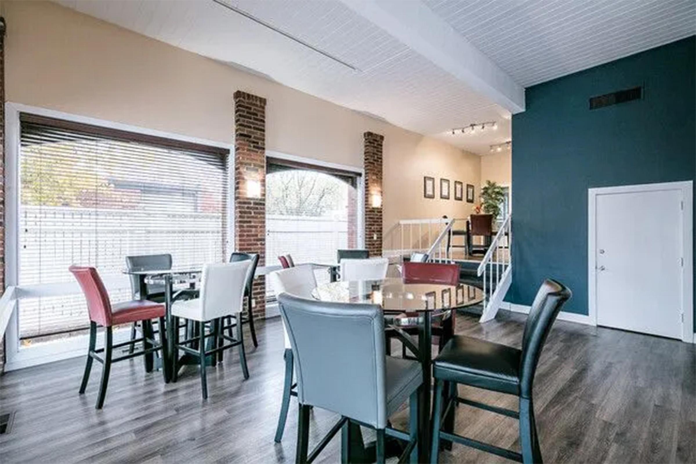 A modern dining area featuring a mix of black, white, and red chairs around tables. Large windows allow natural light to fill the space, highlighting brick columns and hardwood floors. An open staircase leads to an upper level, with decorative plants on the wall.