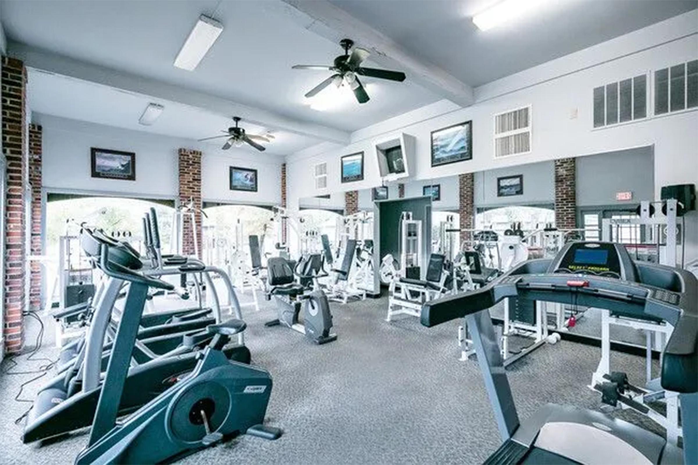 A well-lit gym interior featuring various exercise equipment, including treadmills, stationary bikes, and weight machines. The walls are adorned with framed pictures and there are ceiling fans for ventilation. The space appears clean and organized, inviting for workouts.