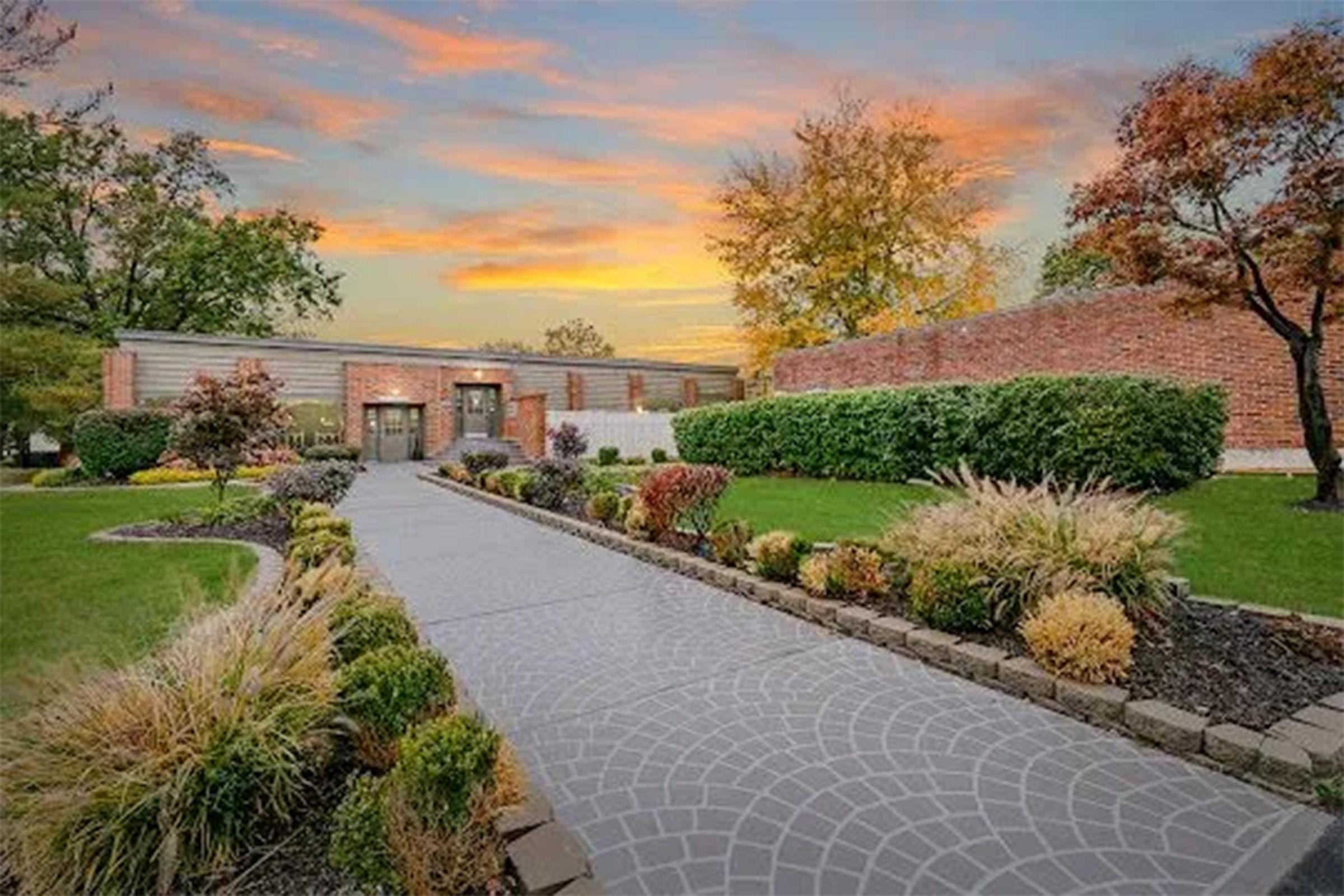 A modern brick house with a landscaped front yard, featuring a stone walkway leading to the entrance. Lush green grass and various shrubs and plants surround the path. In the background, a colorful sunset illuminates the sky, adding a warm glow to the scene.