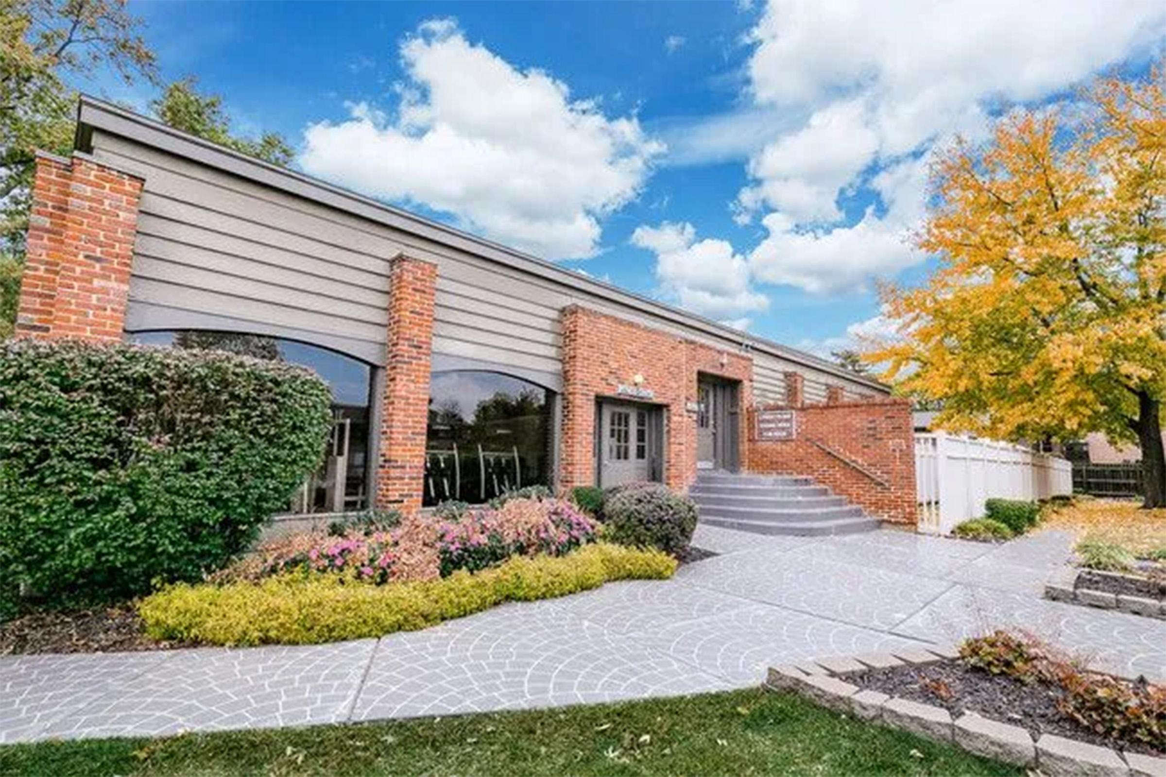A modern brick building with large windows, surrounded by landscaped gardens featuring colorful flowers and shrubs. There are steps leading up to the entrance, and a tree with autumn leaves on the right. The sky is partly cloudy, creating a bright, inviting atmosphere.