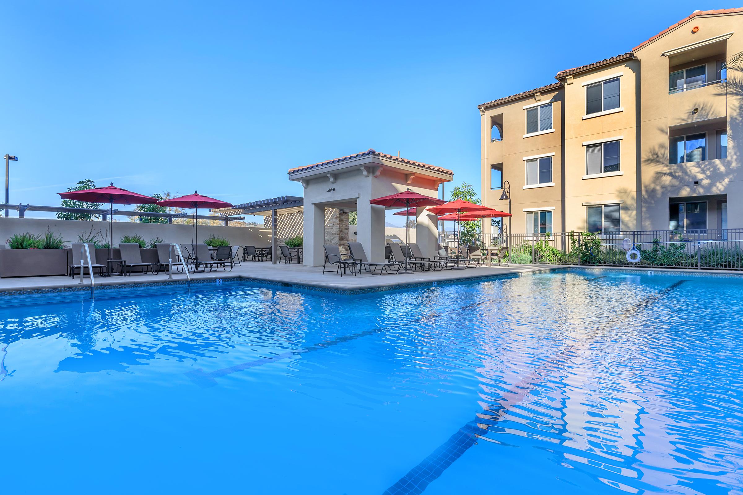 A swimming pool surrounded by lounge chairs and red umbrellas, with a shaded cabana area in the center. The pool is clear and reflects the blue sky. In the background, there is a multi-story building with balconies, adding to the relaxing atmosphere.