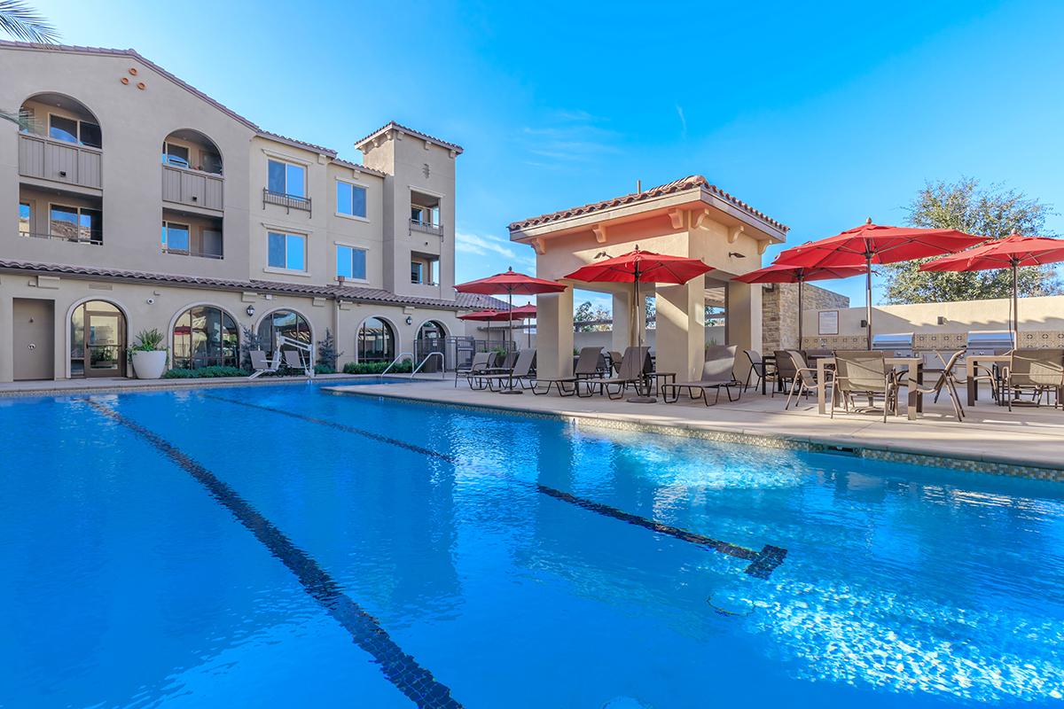 A well-maintained swimming pool surrounded by lounge chairs and red umbrellas, with a multi-story building in the background. The scene is sunny, highlighting the clear blue water and a peaceful atmosphere typical of a resort or apartment complex.