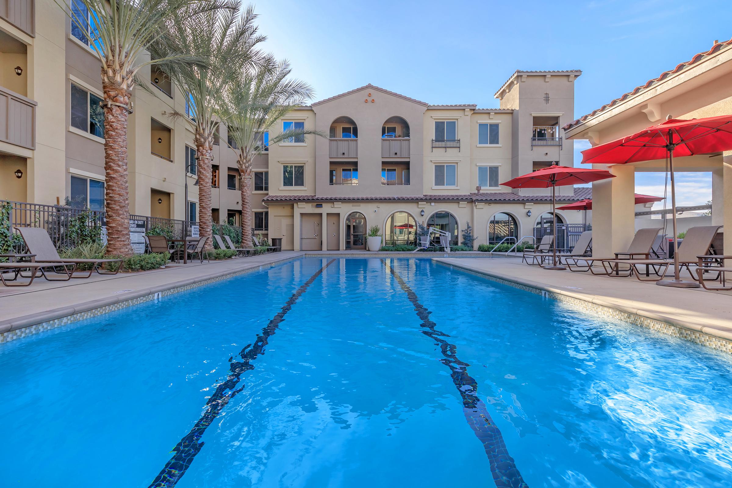 A clear swimming pool surrounded by lounge chairs and red umbrellas, with palm trees in the background. The pool area is part of a residential building that features archways and multiple balconies, under a bright blue sky.