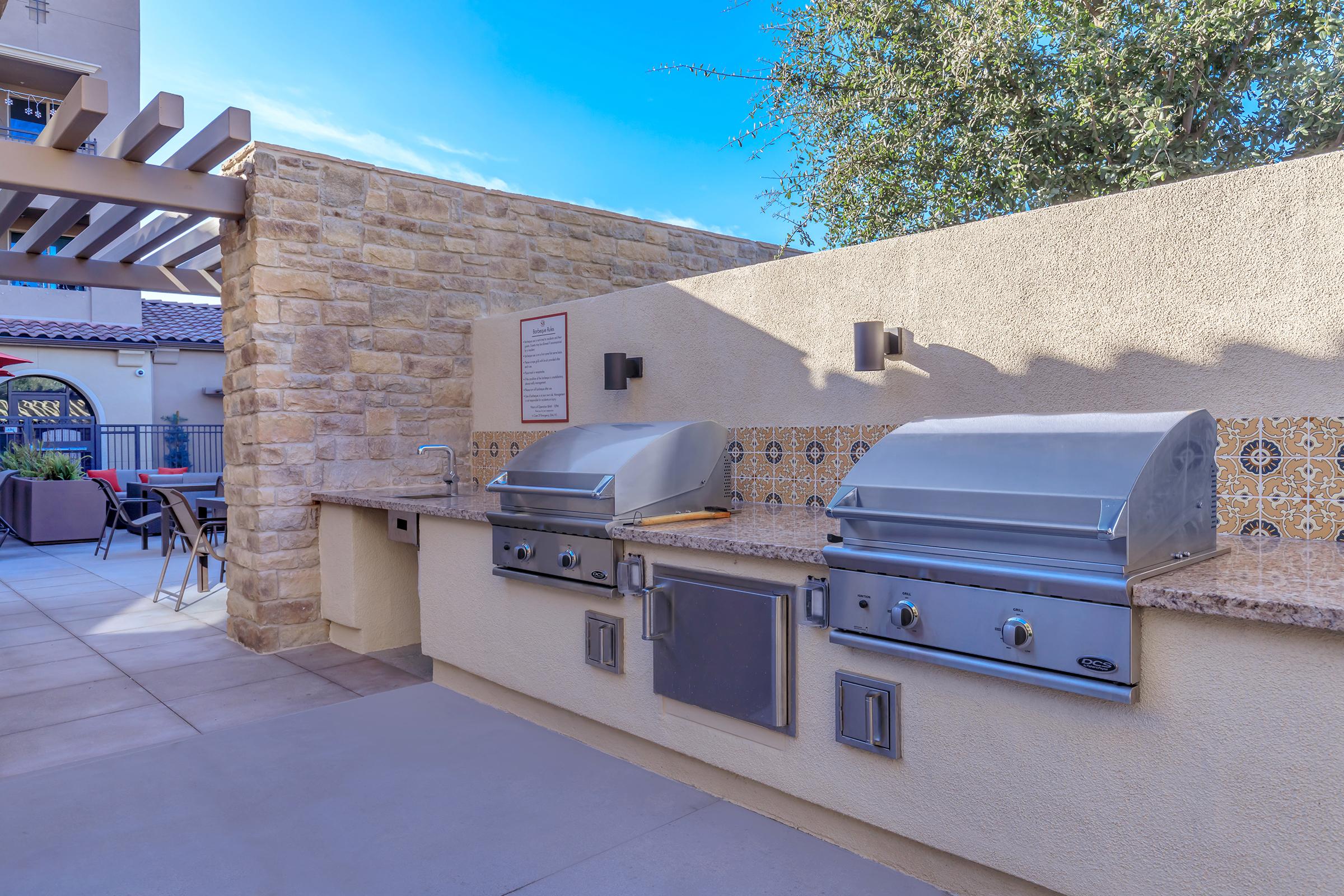 Outdoor grill area featuring two stainless steel barbecue grills on a stone countertop, surrounded by a beige wall with decorative tiles. There's a shaded seating area in the background, and the sky is clear. This space is designed for outdoor cooking and relaxation.