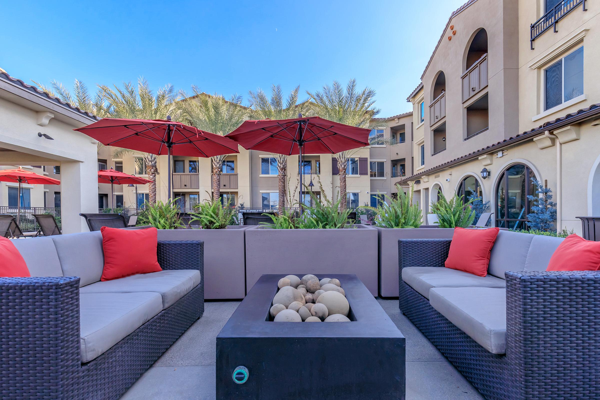 A cozy outdoor lounge area featuring two stylish sofas with red cushions, surrounding a modern fire pit. The space is adorned with palm trees and outdoor umbrellas, set against a backdrop of a multi-story building, creating a welcoming atmosphere for relaxation and social gatherings. Clear blue skies complete the scene.