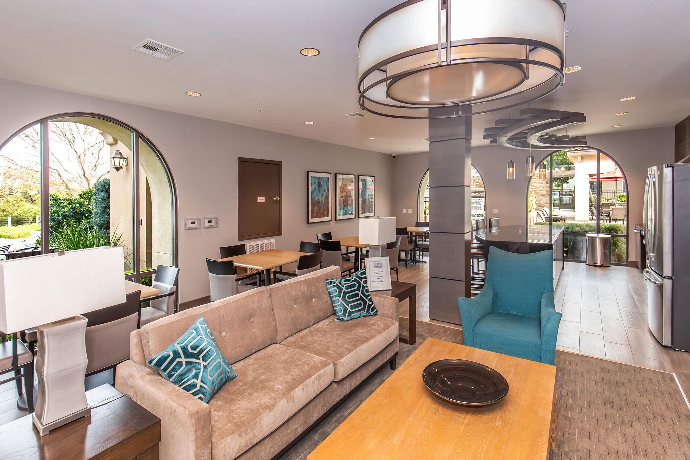 Modern lobby area featuring a comfortable seating arrangement with a sofa and decorative cushions, coffee table, and stylish lighting. The space includes dining tables and chairs, large windows allowing natural light, and contemporary artwork on the walls, creating a welcoming atmosphere.