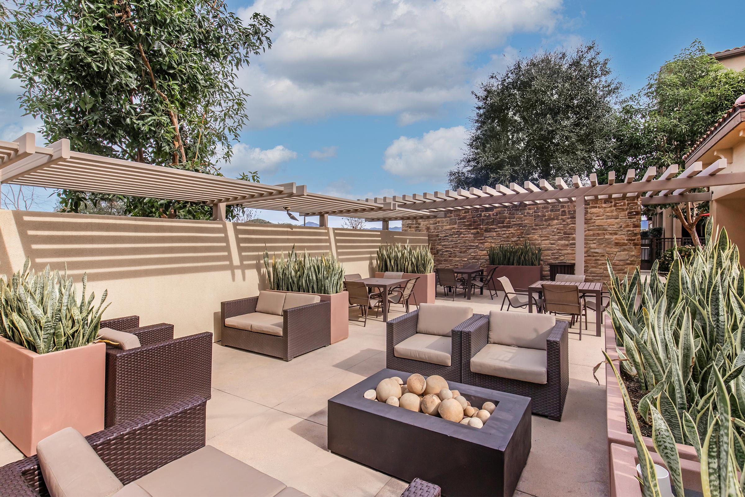 A modern outdoor lounge area featuring comfortable seating with brown wicker chairs and beige cushions, a fire pit with decorative stones at the center, and surrounding greenery. A pergola provides partial shade, and the landscape includes shrubs and trees under a partly cloudy blue sky.