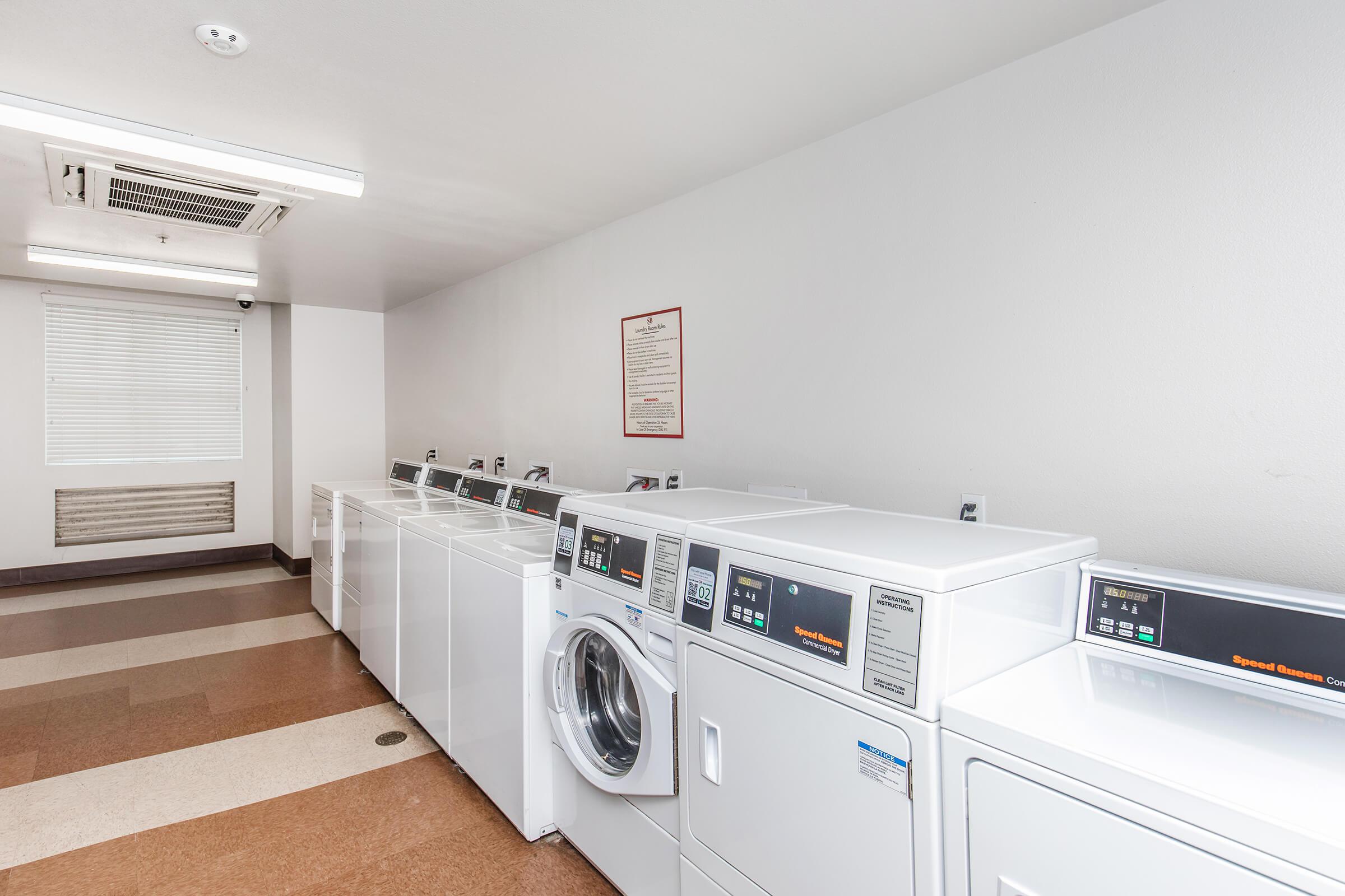 Laundry room with several white washing machines and dryers lined up against a wall. A window with blinds is partially open, providing natural light. There is a sign on the wall with instructions or information. The floor features a combination of brown and beige tiles.