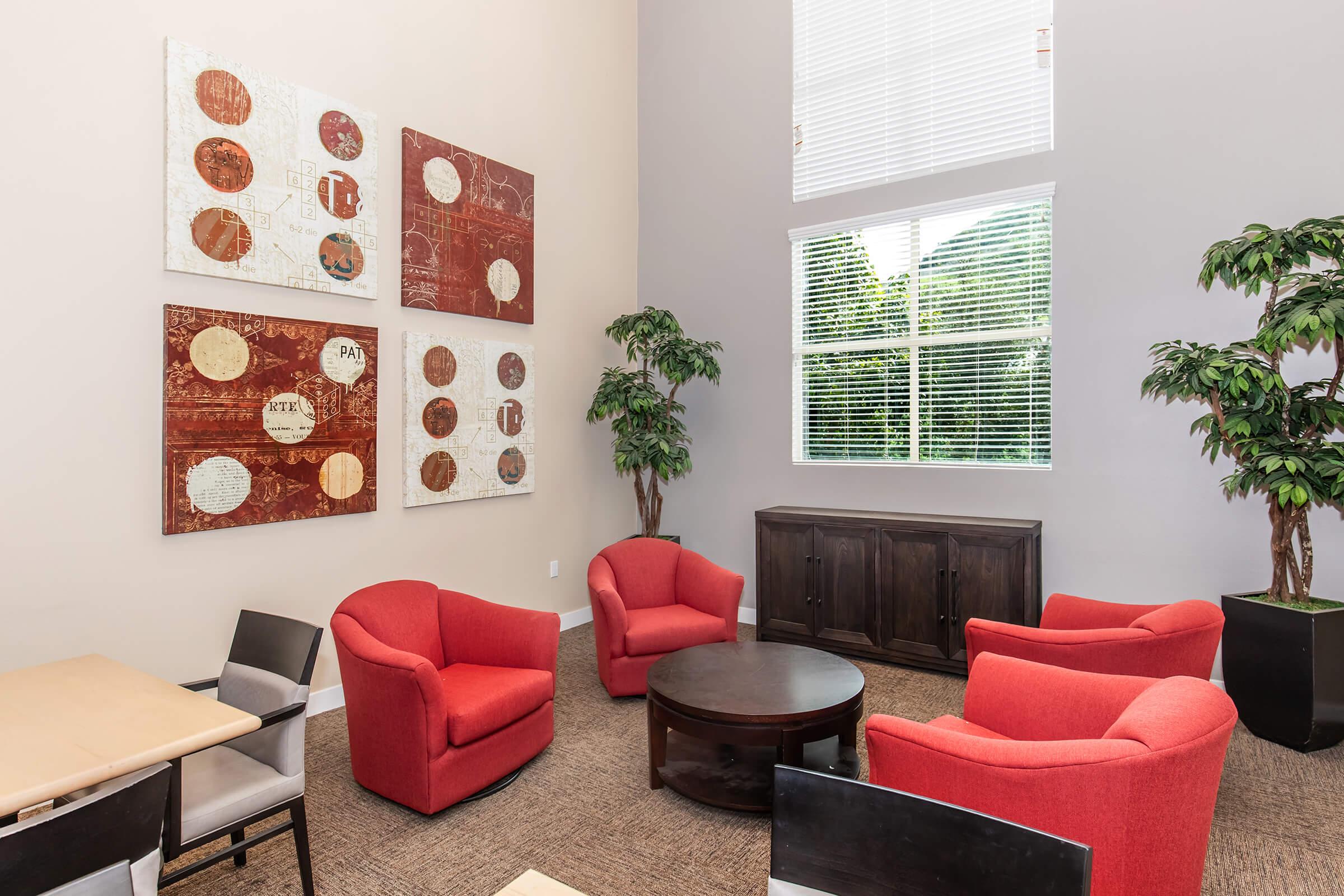 A cozy lounge area featuring four red armchairs arranged around a circular table. The walls are adorned with abstract art pieces in warm tones. A wooden cabinet is positioned against one wall, and large windows allow natural light to illuminate the space, complemented by decorative plants.