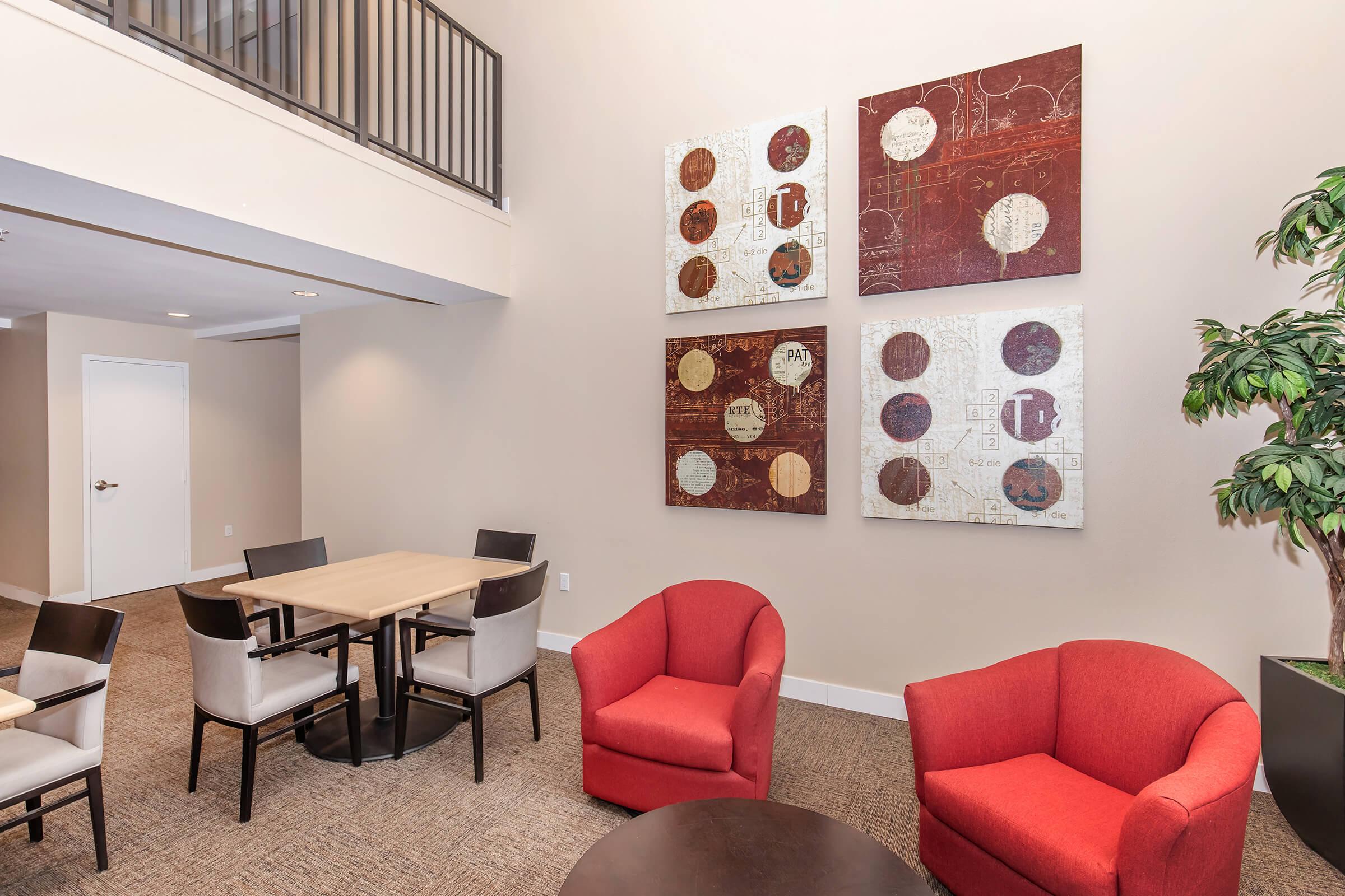 A modern indoor lounge area featuring two round red chairs and tables with four chairs around them. The walls display four abstract artworks with circular designs in earthy tones. A tall indoor plant is positioned near the seating area. The overall decor is contemporary and inviting.