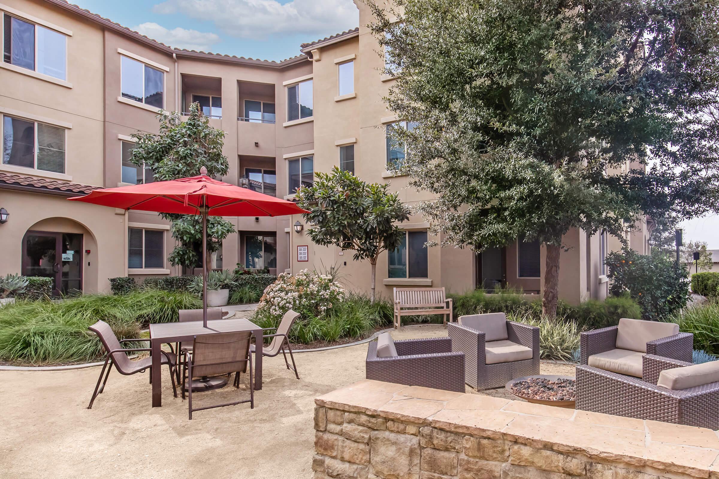 A landscaped courtyard featuring a round table with chairs under a red umbrella. There are several seating arrangements with modern outdoor furniture, surrounded by greenery and landscaping. The backdrop includes an apartment building with multiple windows.