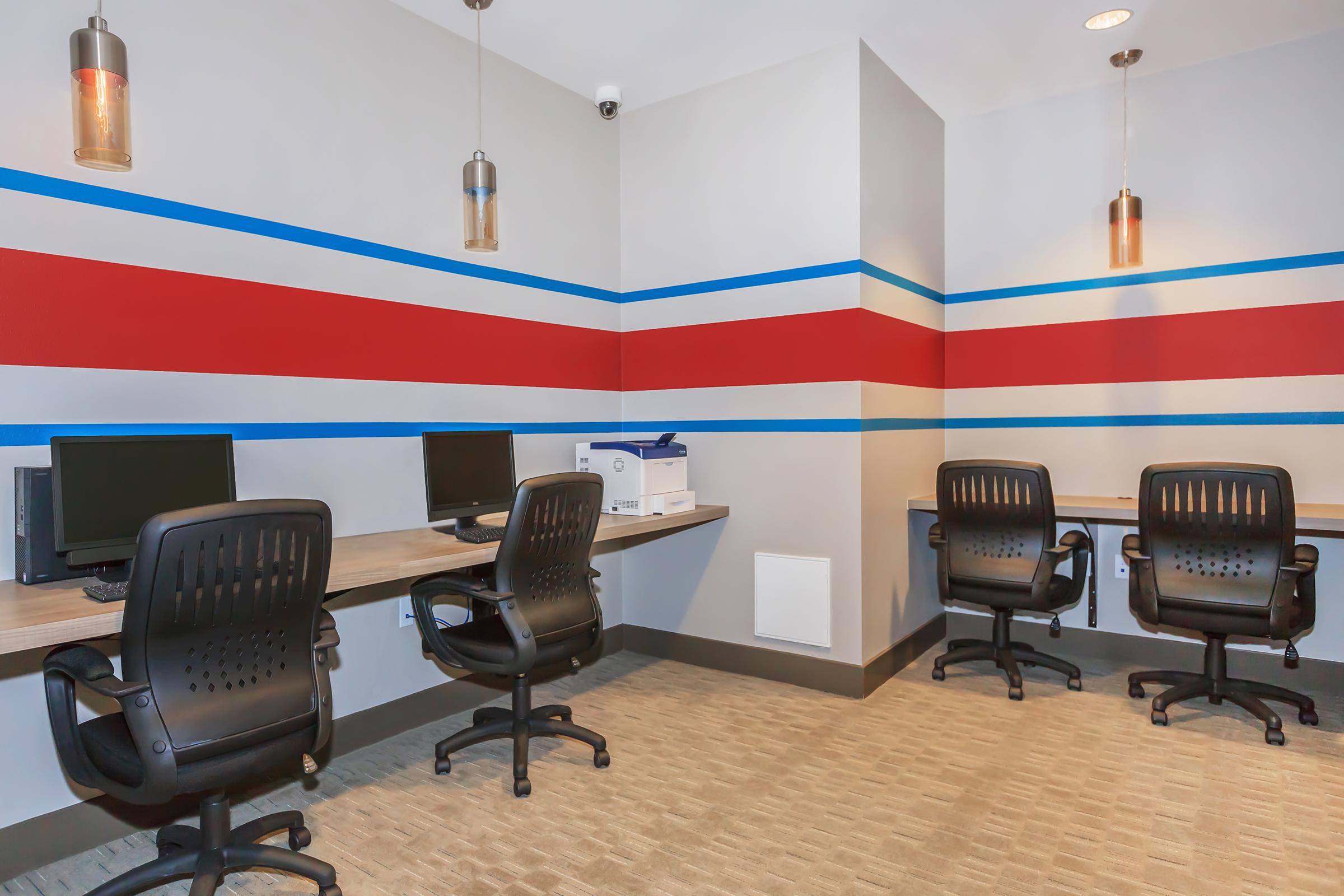 A modern workspace featuring three ergonomic black office chairs and two computers on wooden desks against a wall painted with red and blue stripes. The room is well-lit with pendant lighting and includes a printer on a side table.