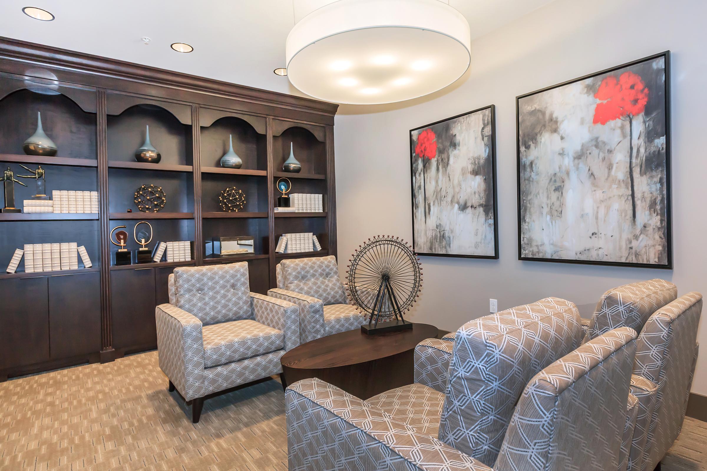 Cozy seating area featuring two patterned armchairs and a round table, set against a backdrop of dark wood shelves filled with decorative items and books. The walls display two large framed artworks with red flowers. Bright overhead lighting enhances the inviting atmosphere.