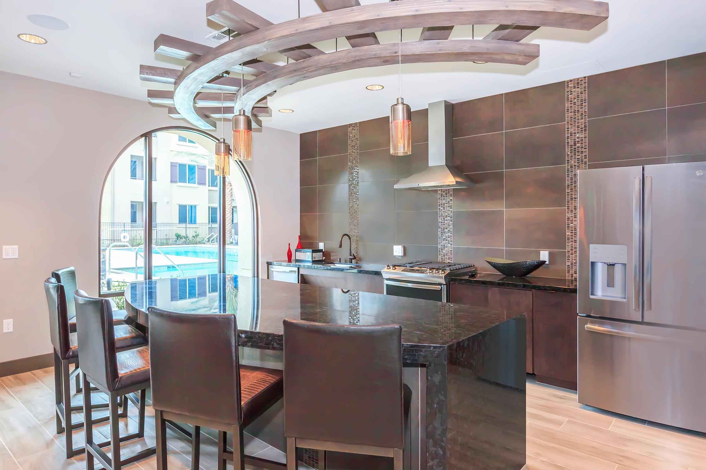 Modern kitchen featuring a large island with dark granite countertops and bar seating. Above the island, a distinctive circular wooden light fixture hangs, complemented by stylish pendant lights. The sleek cabinetry and stainless steel appliances, including a refrigerator and stove, enhance the contemporary design, with a pool view visible through an arched window.