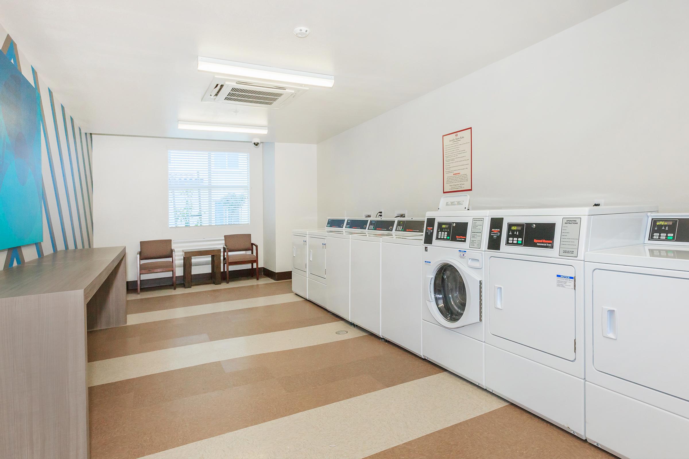 A clean and modern laundry room featuring several white washing machines and dryers lined up against a wall. There are two brown chairs near a window, and the floor is a combination of light and dark beige tiles. An air conditioning unit is visible on the ceiling, and a decorative blue artwork is on the wall.