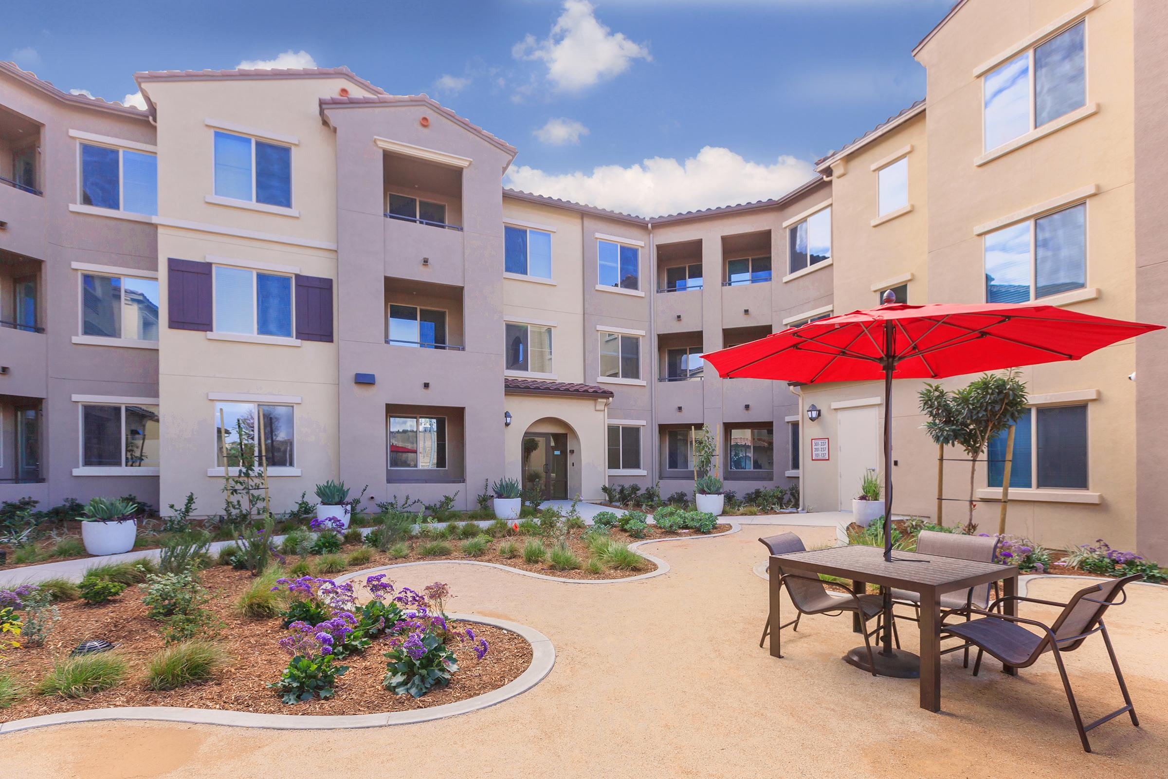 A well-kept courtyard of an apartment building featuring a large red umbrella over a dining table with chairs. Surrounding the table are landscaped areas with flowers and plants, and the building has multiple windows and light-colored walls under a blue sky.