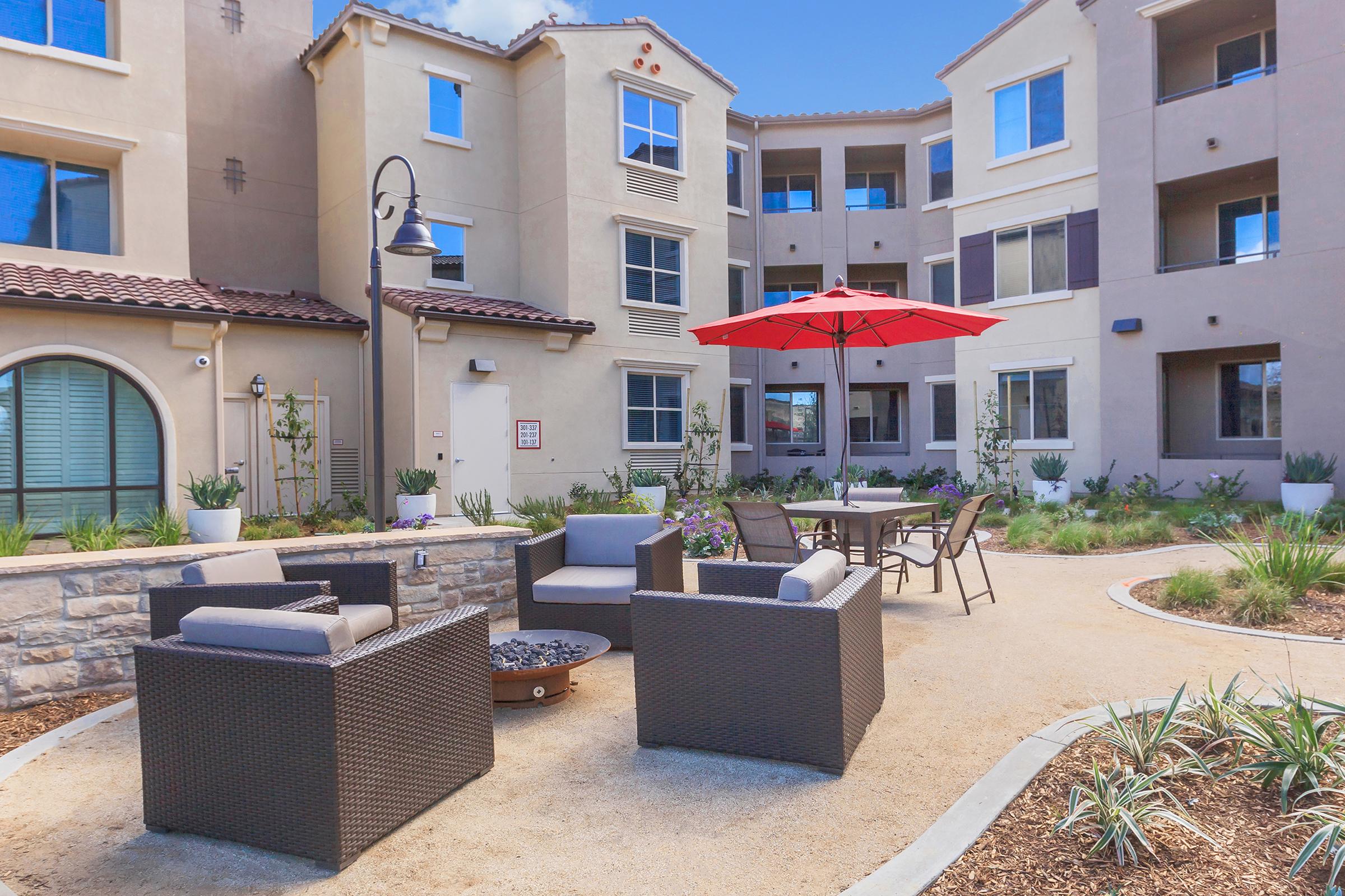 A landscaped courtyard featuring comfortable seating areas with wicker chairs and a round table under a bright red umbrella. The setting is surrounded by modern apartment buildings, with decorative plants and stone elements highlighting the outdoor space. Bright blue sky adds a cheerful ambiance.