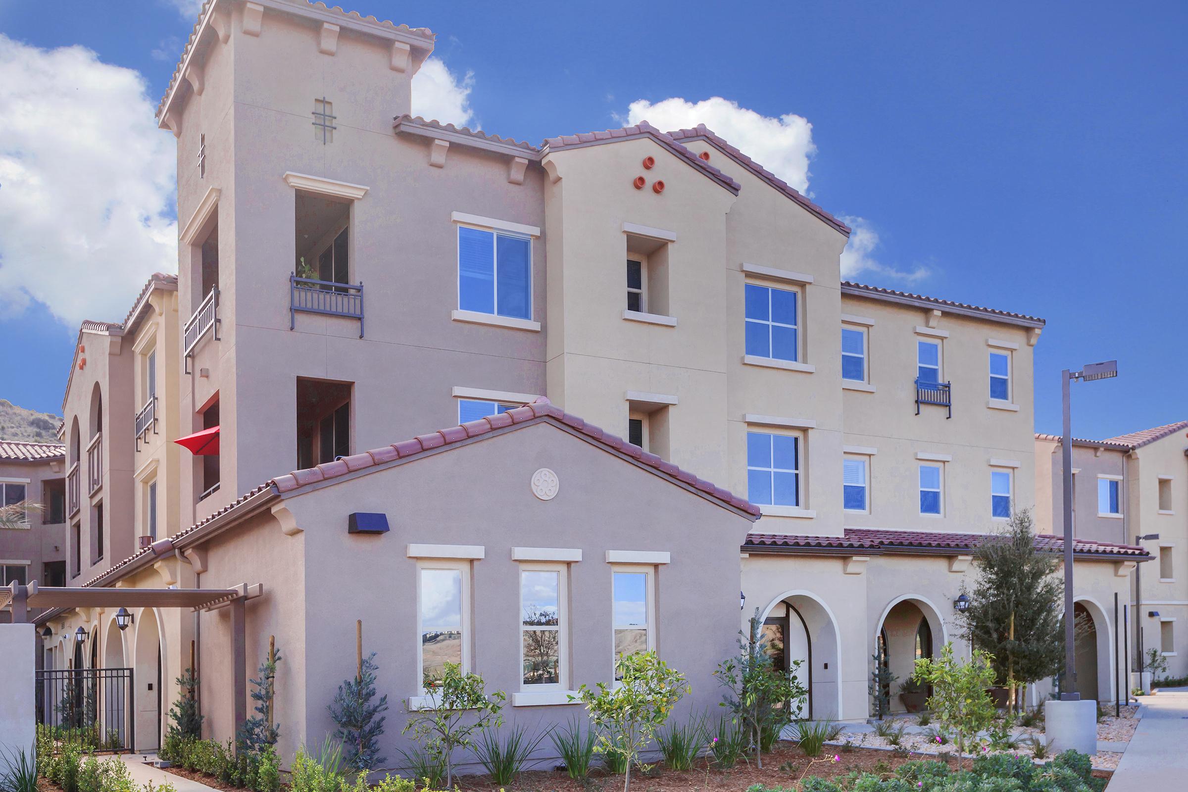 A modern, three-story building with a stucco exterior and a tiled roof. The structure features large windows, balconies, and decorative elements. Lush landscaping is visible in front, with small trees and plants. The background includes a clear blue sky and distant hills, giving a pleasant residential atmosphere.