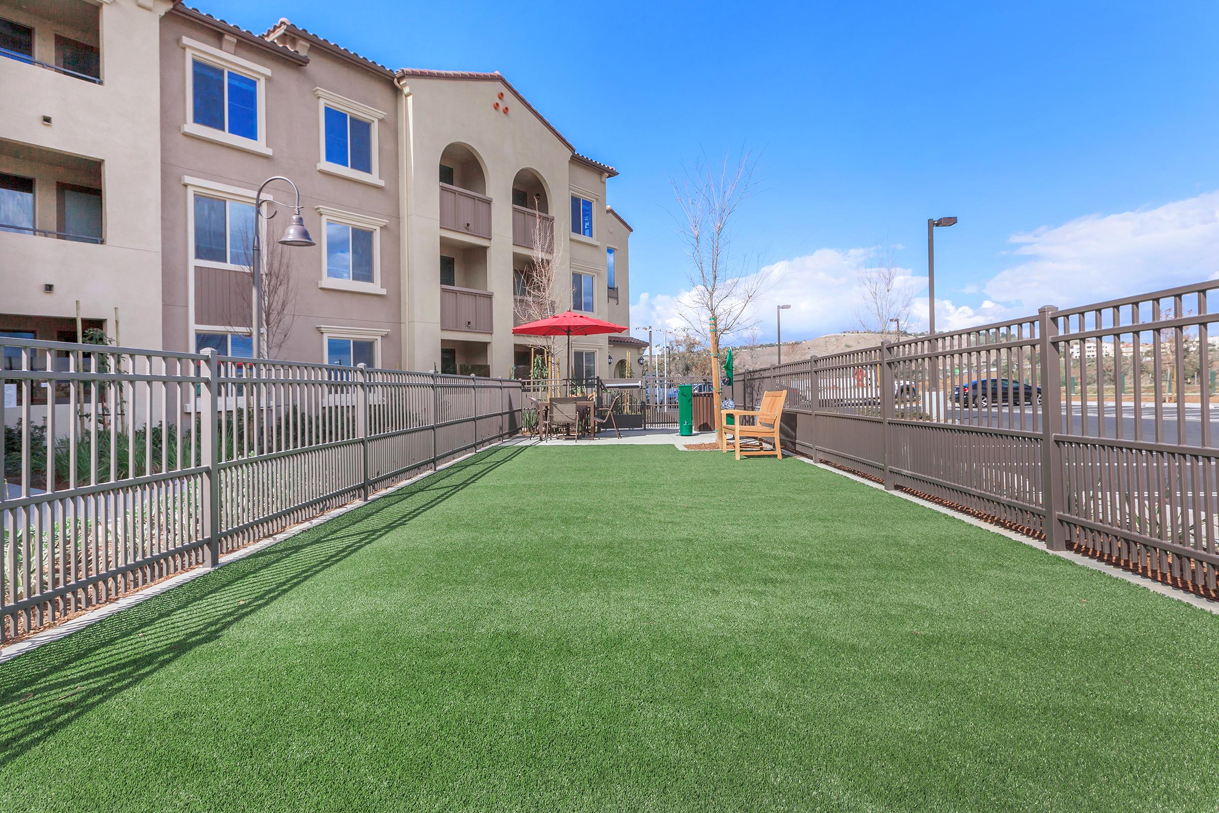 View of a fenced outdoor area with artificial turf, featuring a bench and an umbrella table. In the background, there are residential buildings and clear blue skies. The space offers a welcoming atmosphere for relaxation and outdoor activities.