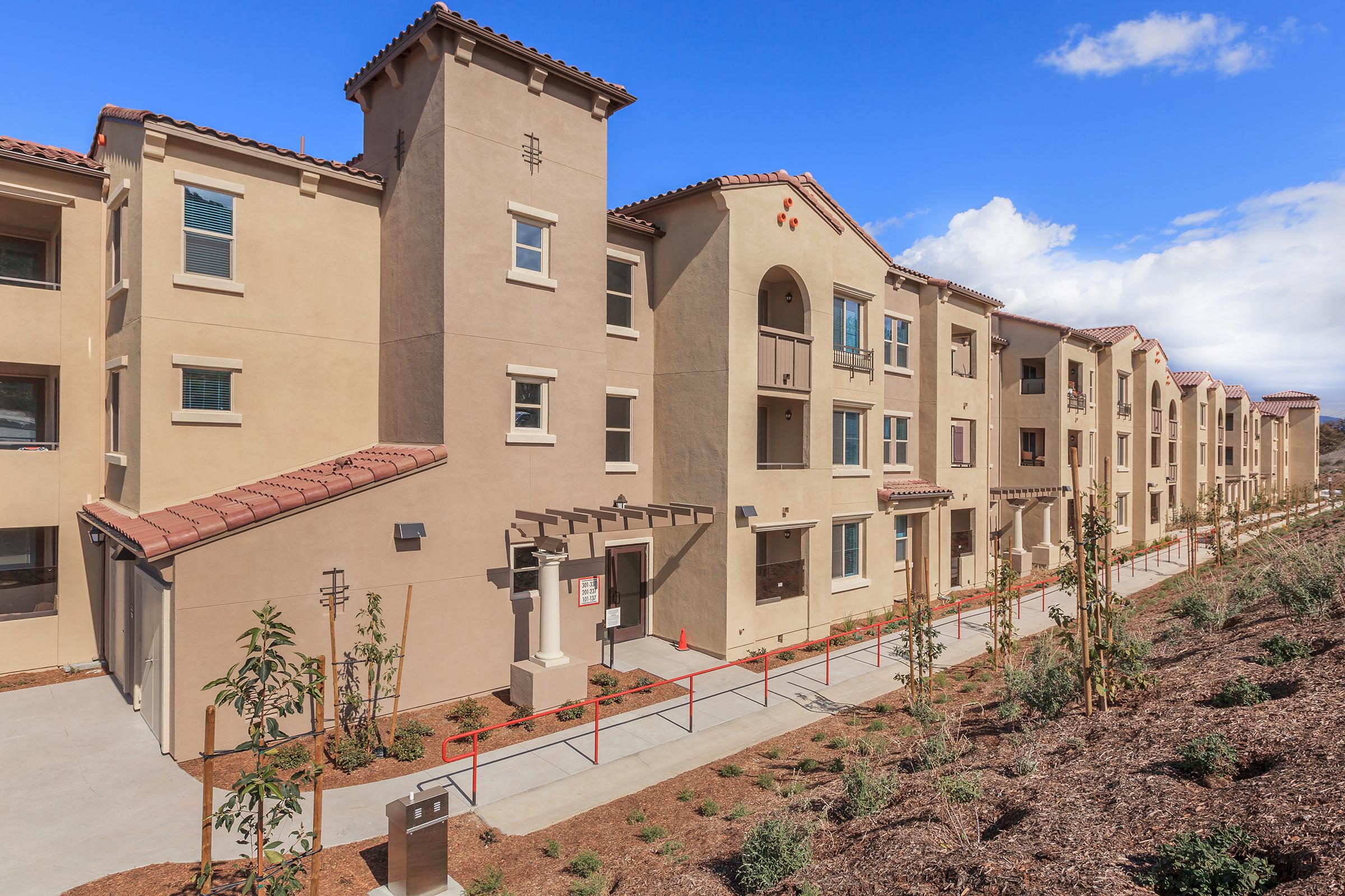A row of modern residential buildings with stucco exteriors and terracotta roofs. The buildings feature multiple windows and balconies, along with a landscaped area in front. Sidewalks and pathways are lined with young trees and shrubs, under a partly cloudy sky.