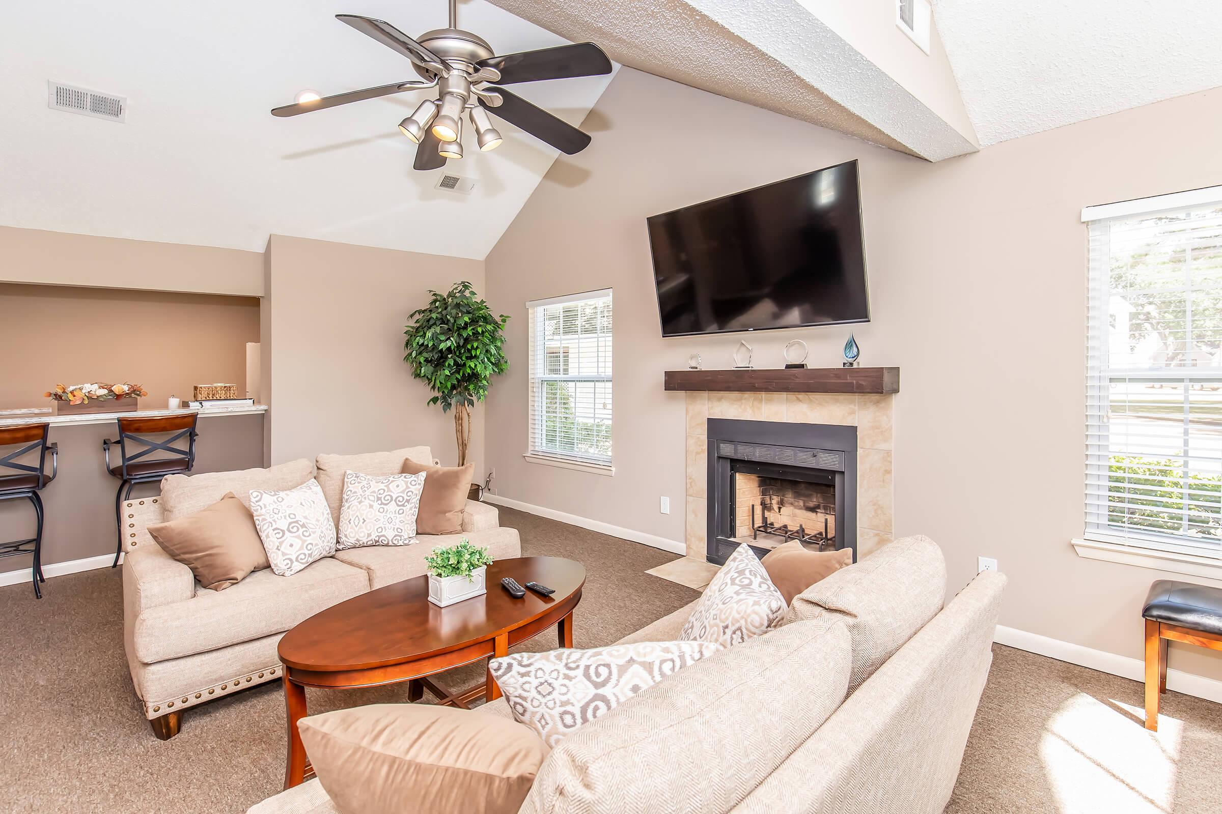 A modern living room featuring a beige sofa with decorative pillows, a coffee table, and a ceiling fan. A mounted flat-screen TV is on the wall above a cozy fireplace. Natural light enters through large windows, creating a warm and inviting atmosphere. A potted plant adds a touch of greenery.