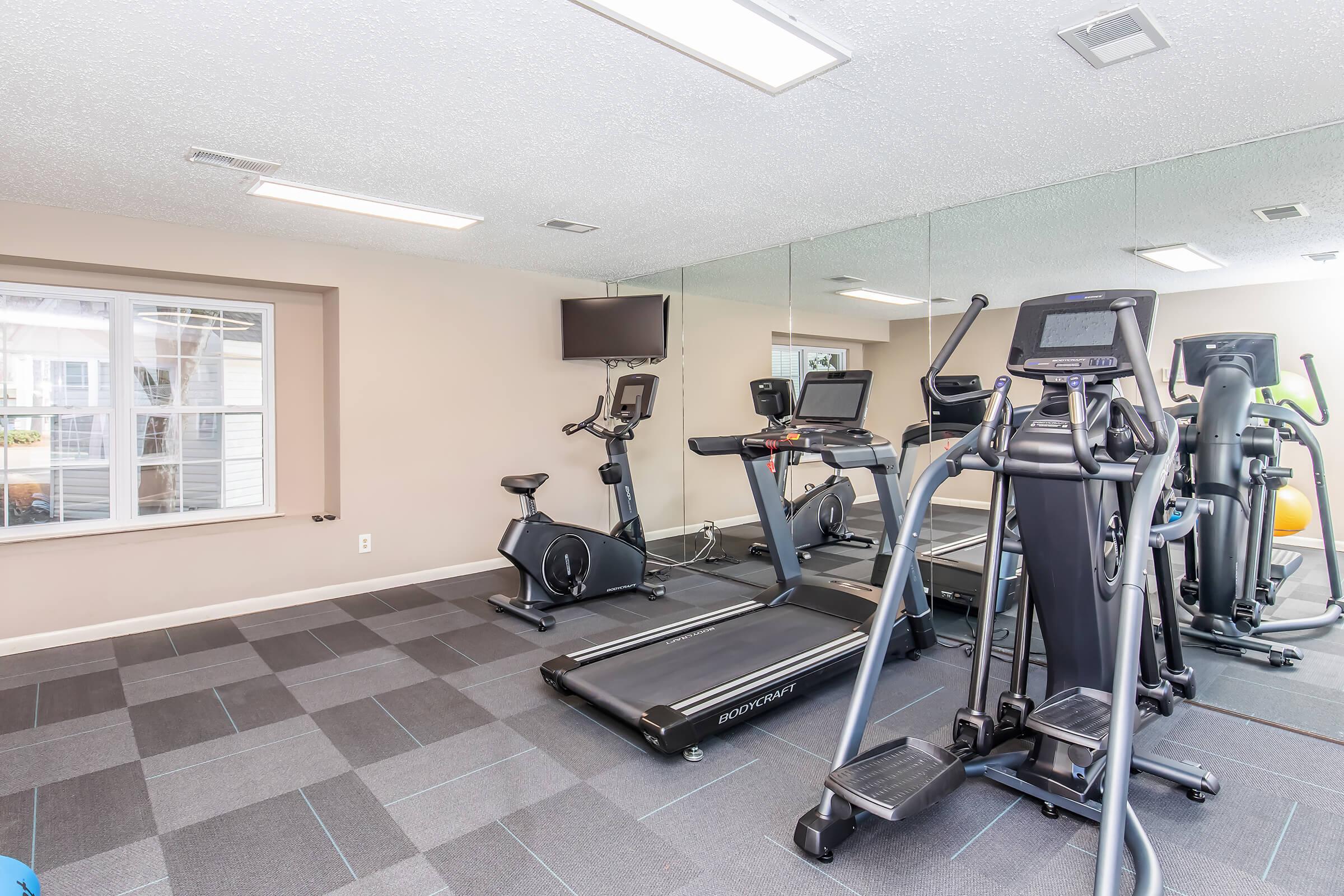 A well-equipped gym with a treadmill, elliptical machine, and stationary bike. The room features mirrors, a television on the wall, and large windows allowing natural light to fill the space. The flooring is a modern, dark tile with a patterned design.