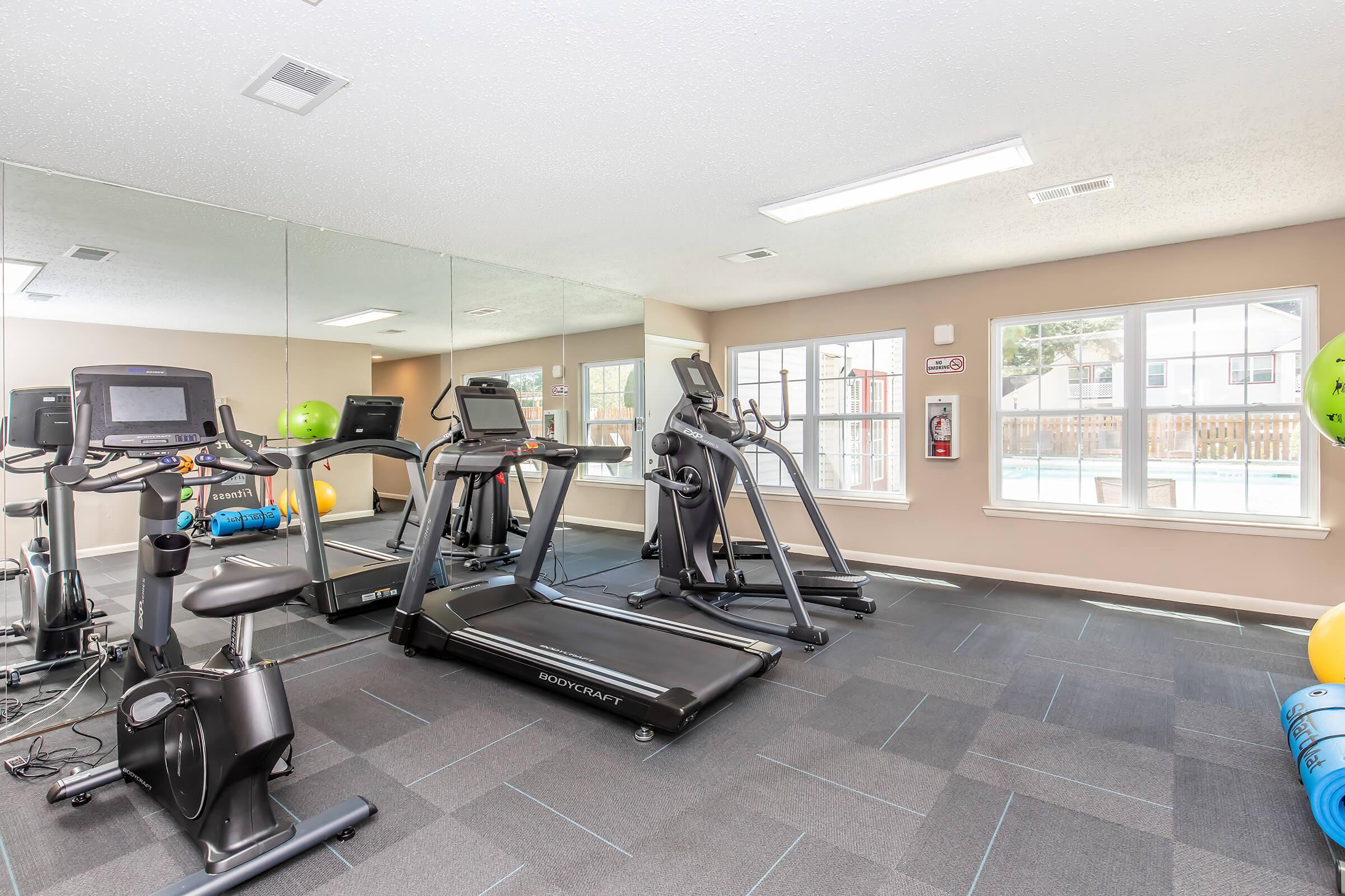 A bright fitness room featuring exercise equipment, including a treadmill, stationary bike, and elliptical machine, set against a mirrored wall. Large windows provide natural light and a view of an outdoor pool area, with colorful exercise balls in the corner.