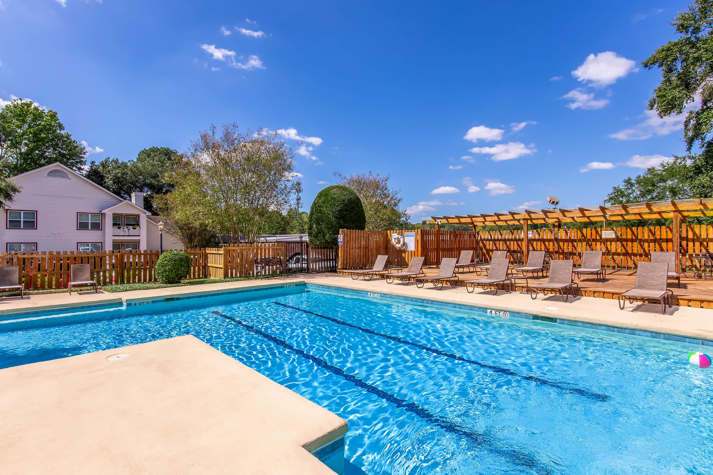 A clear blue swimming pool surrounded by lounge chairs and wooden decking. In the background, there are trees and a wooden fence. The sky is bright and partly cloudy, creating a sunny atmosphere.