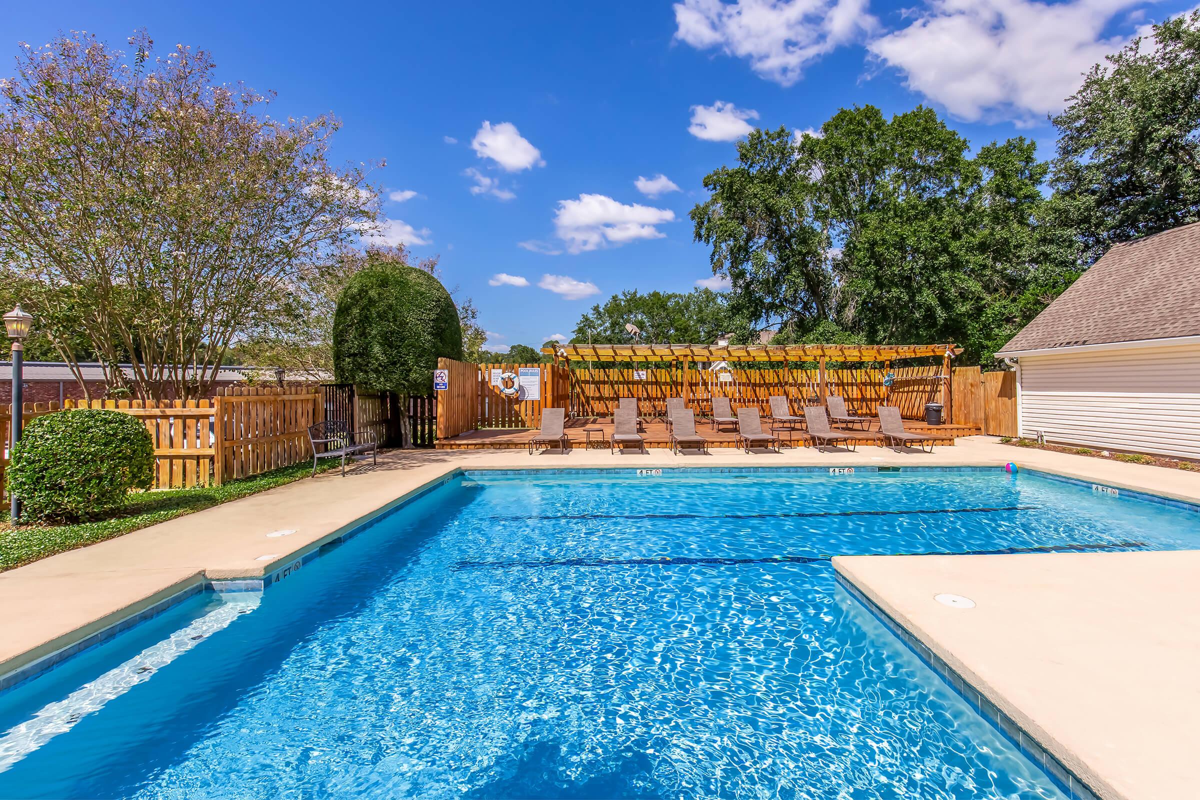 A sunny outdoor swimming pool surrounded by wooden deck chairs, with a fenced area and trees in the background. The pool is clear and inviting, under a bright blue sky with a few fluffy clouds.