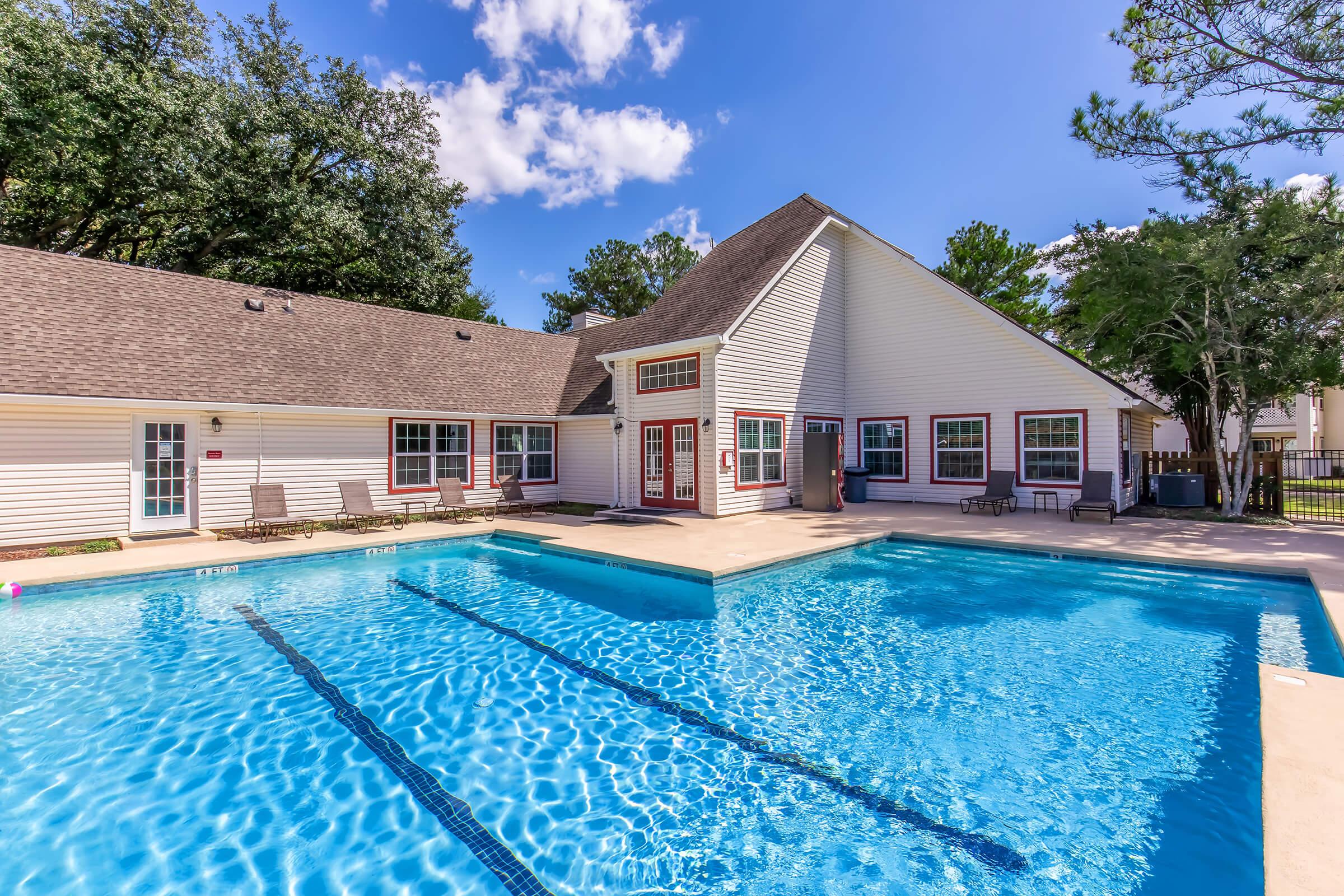 A bright, sunny outdoor pool area featuring two swimming pools with clear blue water. Surrounding the pools are lounge chairs and a building with a sloped roof, large windows, and a shaded porch. Trees are in the background under a blue sky with scattered clouds.