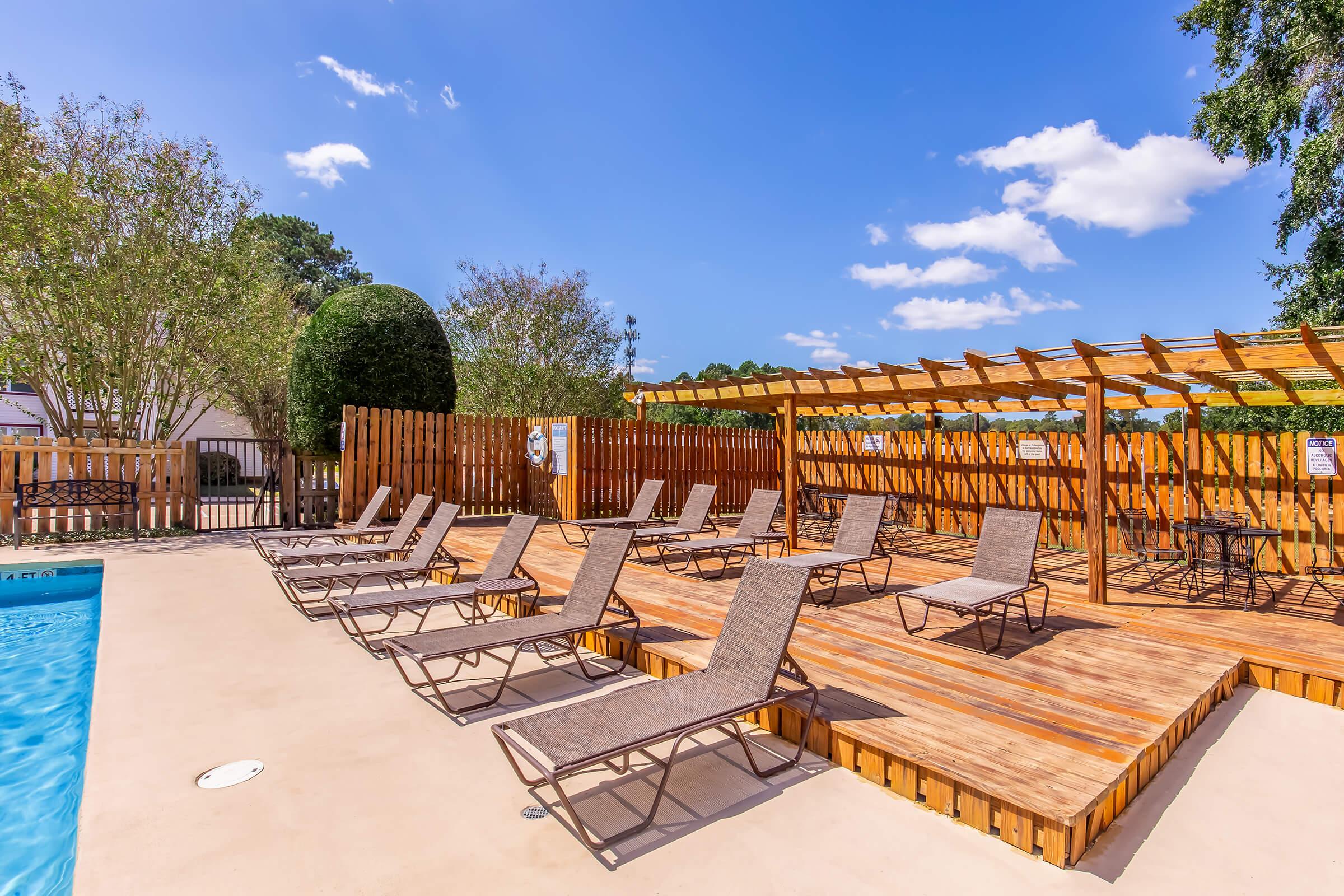 A sunlit outdoor pool area featuring a row of lounge chairs arranged on a wooden deck. In the background, there are trees and a wooden fence with shaded pergolas. The sky is clear with a few clouds, creating a relaxing atmosphere perfect for sunbathing and leisure.