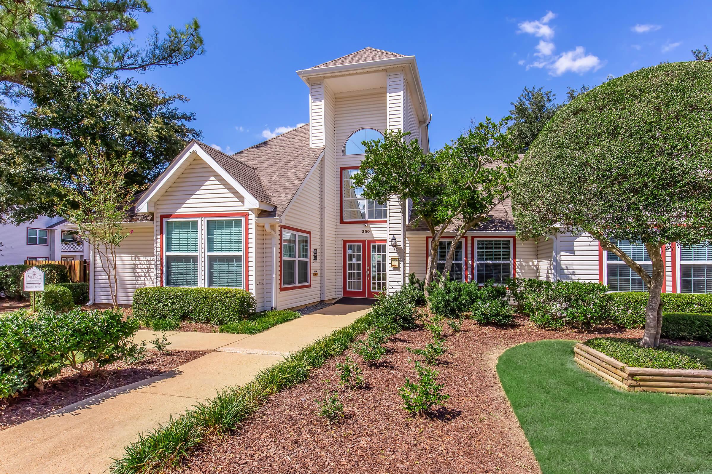 A well-maintained residential building with a large entrance featuring a tall central tower. The exterior has a mix of white and red trim, surrounded by neatly landscaped gardens with shrubs and ornamental trees. A pathway leads up to the entrance, with blue skies and scattered clouds above.