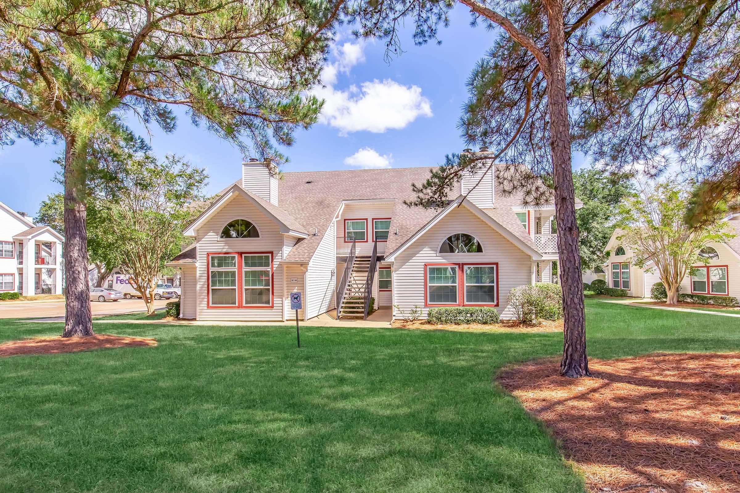 Two-story residential building with a light-colored exterior, large windows, and a welcoming porch. Surrounded by green grass and several trees, under a bright blue sky with fluffy white clouds. Nearby pathways and other buildings in a well-maintained community setting.
