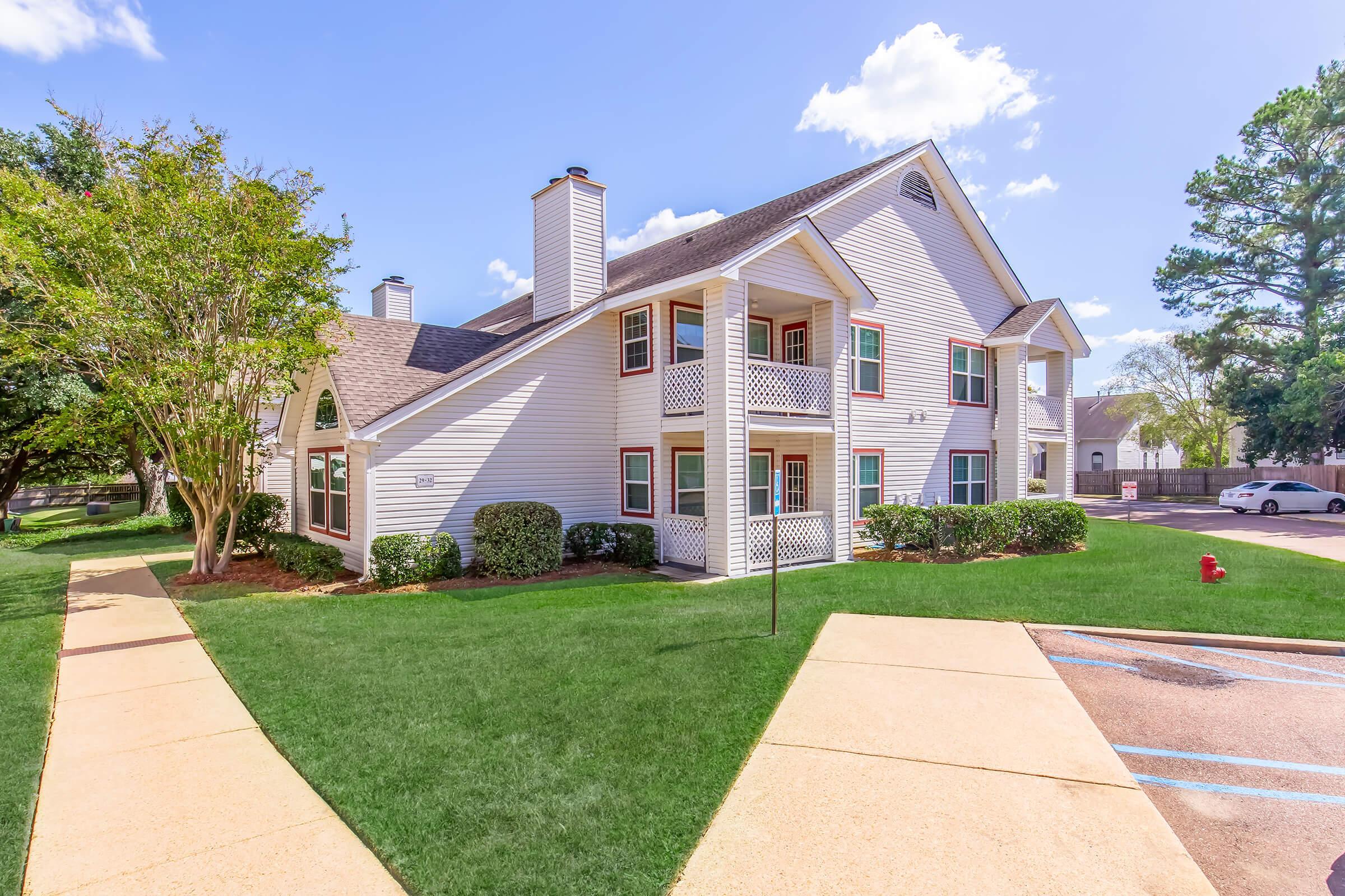 A two-story residential building with white siding and dark roofing, surrounded by green grass and small shrubs. There are sidewalks leading to the front entrance, and a bright blue sky with a few clouds overhead. Trees are visible in the background, creating a serene neighborhood atmosphere.