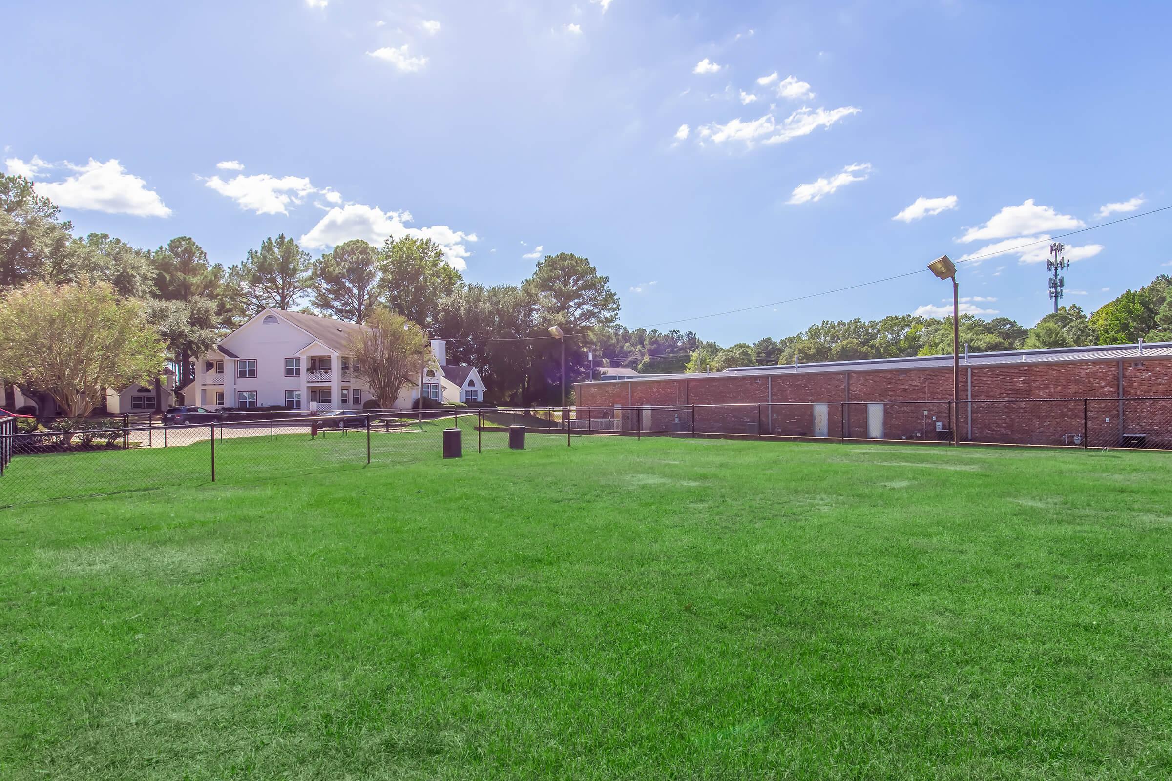 A spacious green field surrounded by a fence, with a few trees in the background. In the distance, there are two buildings: one is a white multi-story structure and the other is a low brick building. The sky is clear with a few clouds.