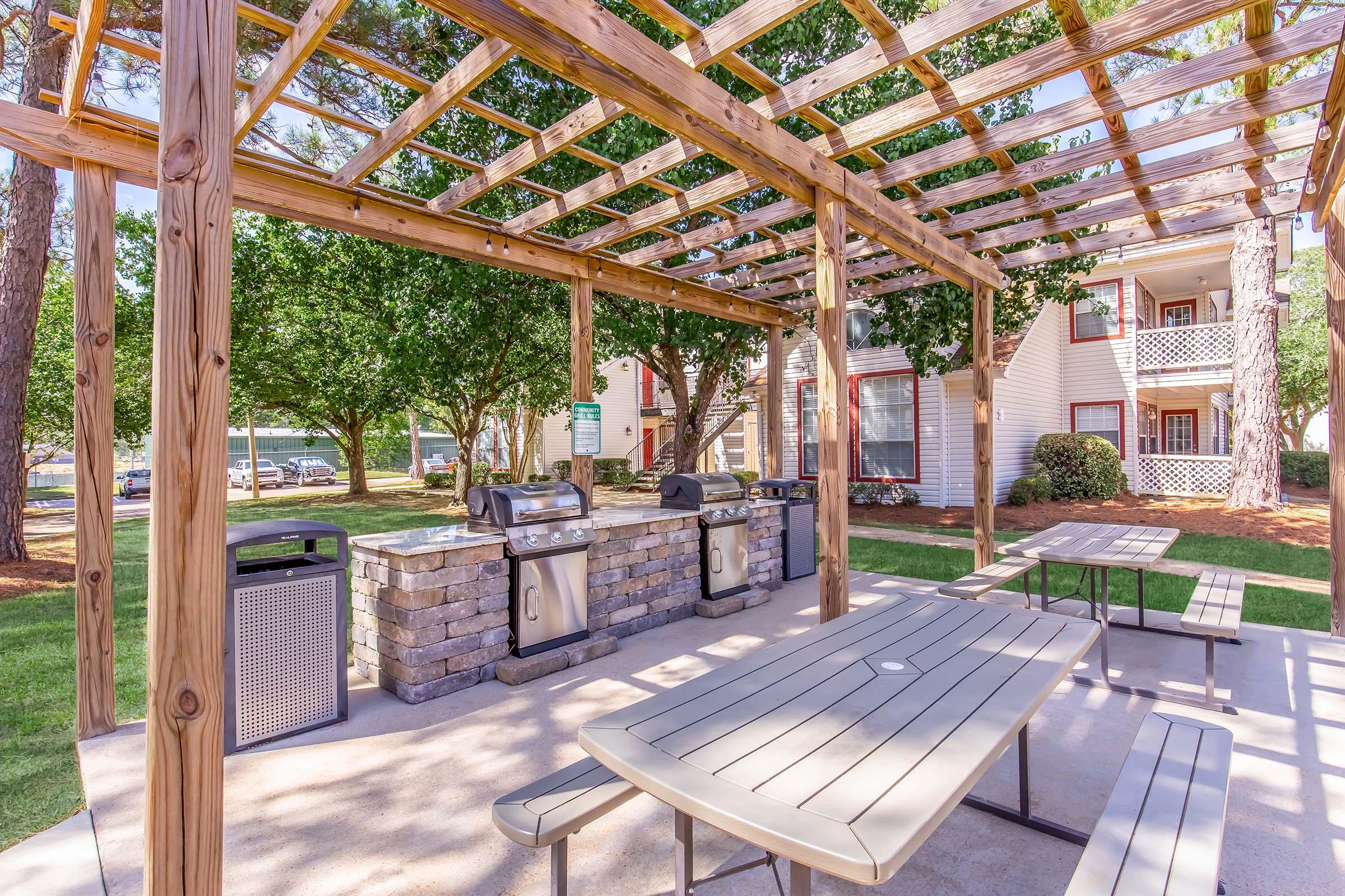 Outdoor picnic area with a pergola, featuring several grills and a stone countertop. There are tables for seating underneath the structure, surrounded by green trees and nearby residential buildings. The area is designed for social gatherings and cooking.