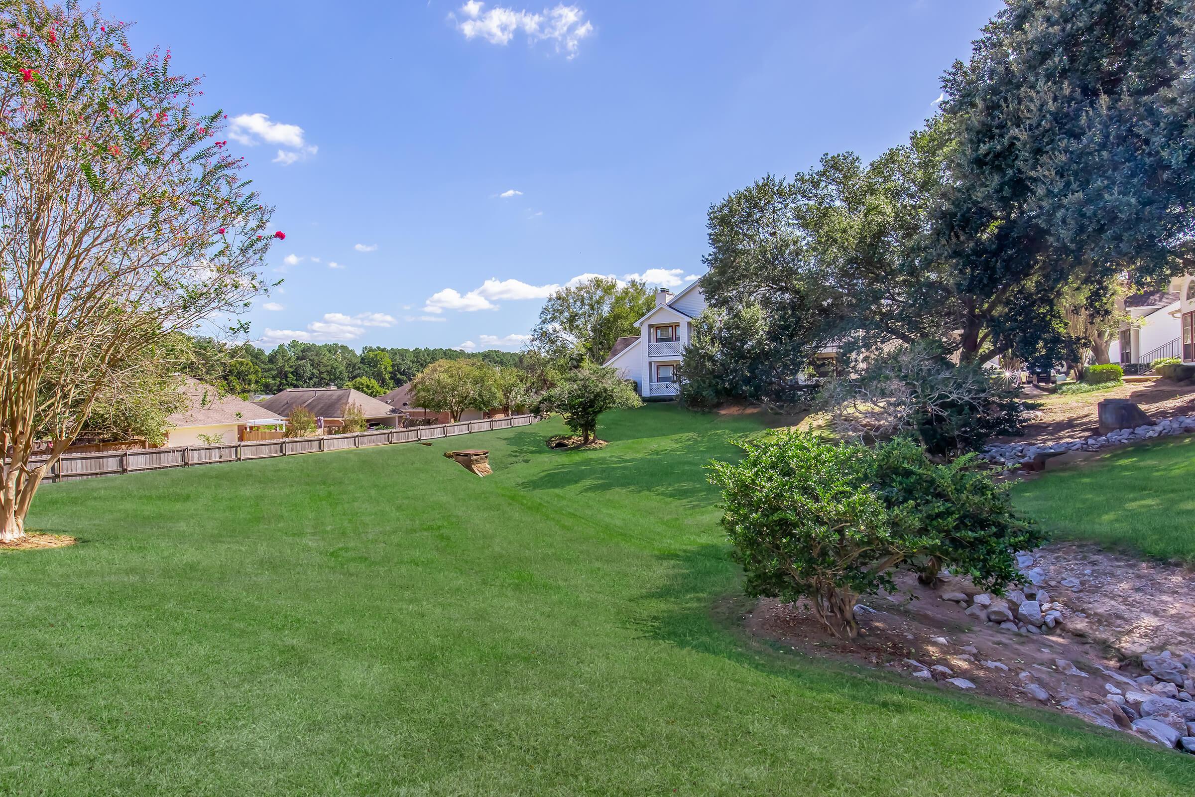 A spacious, well-maintained lawn with lush green grass under a bright blue sky. Trees line the property, and a small rock area is visible to the right. In the background, a white house is partially obscured by trees, and residential homes can be seen beyond the fence. The scene is calm and inviting.