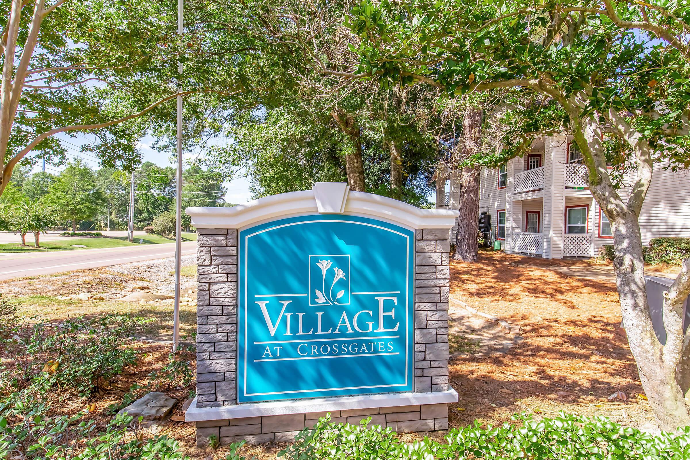 Sign for "Village at Crossgates" featuring a blue background with white lettering, surrounded by greenery and trees. The sign has a stone base and is located near a road.