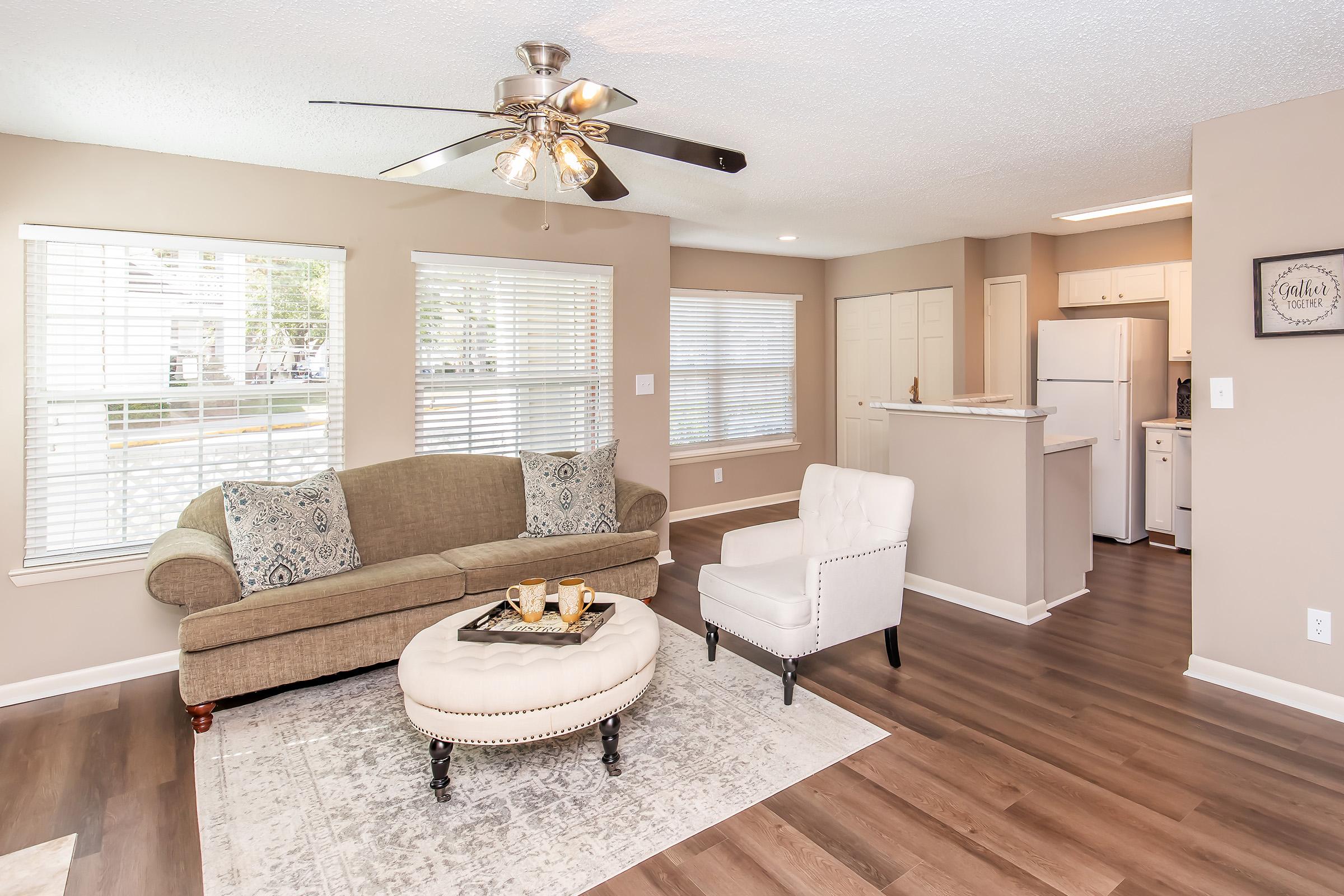 A cozy living room featuring a light brown sofa with decorative pillows, a round coffee table with drinks, and a white armchair. The space has large windows with blinds, allowing natural light to fill the room. A ceiling fan hangs above, and the floor has modern wood-like laminate. The kitchen area is visible in the background.