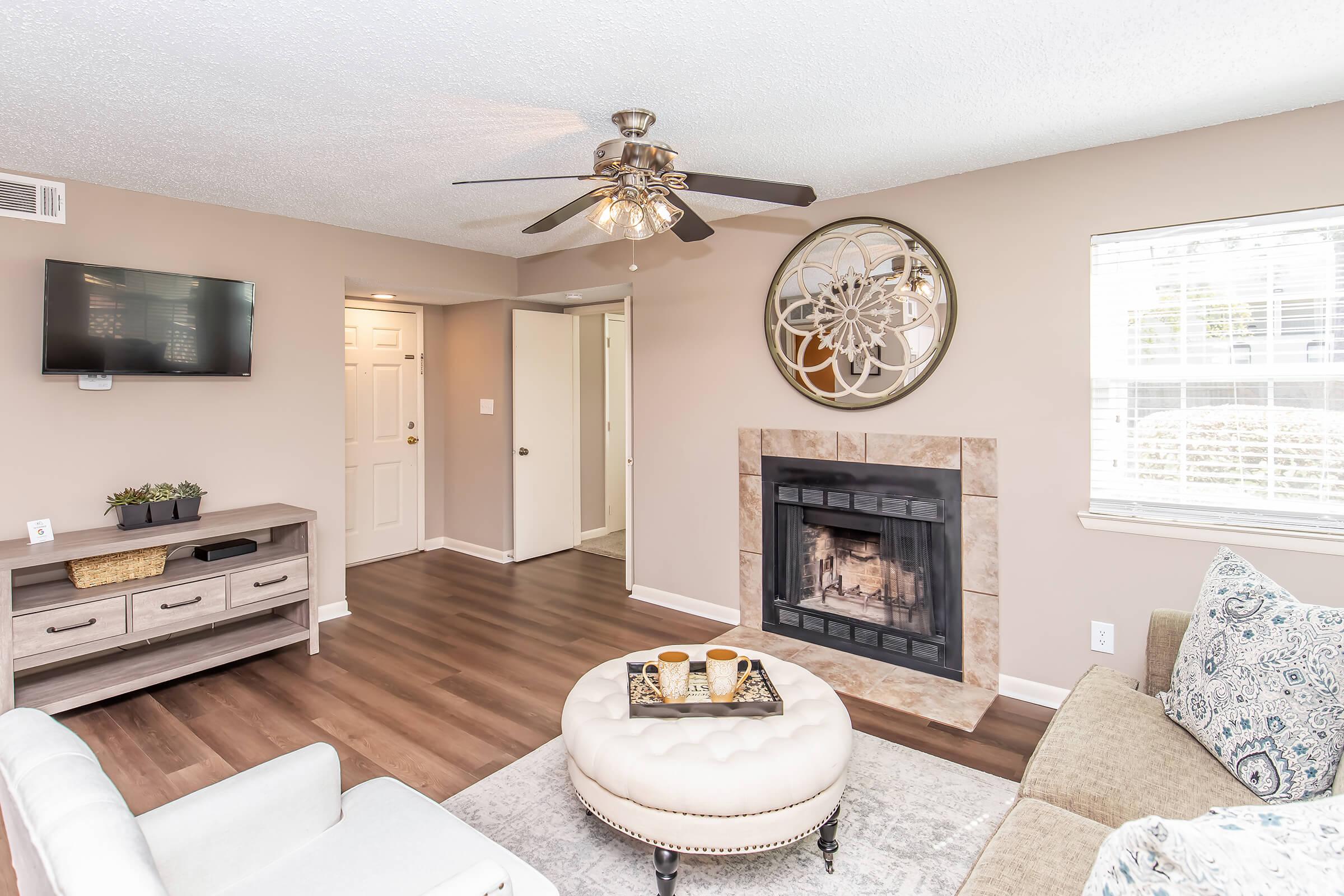 Cozy living room featuring a ceiling fan, modern furniture including a light-colored sofa and an ottoman, a flat-screen TV mounted on the wall, and a decorative wall clock. A fireplace adds warmth to the space, complemented by soft lighting and neutral-toned walls.