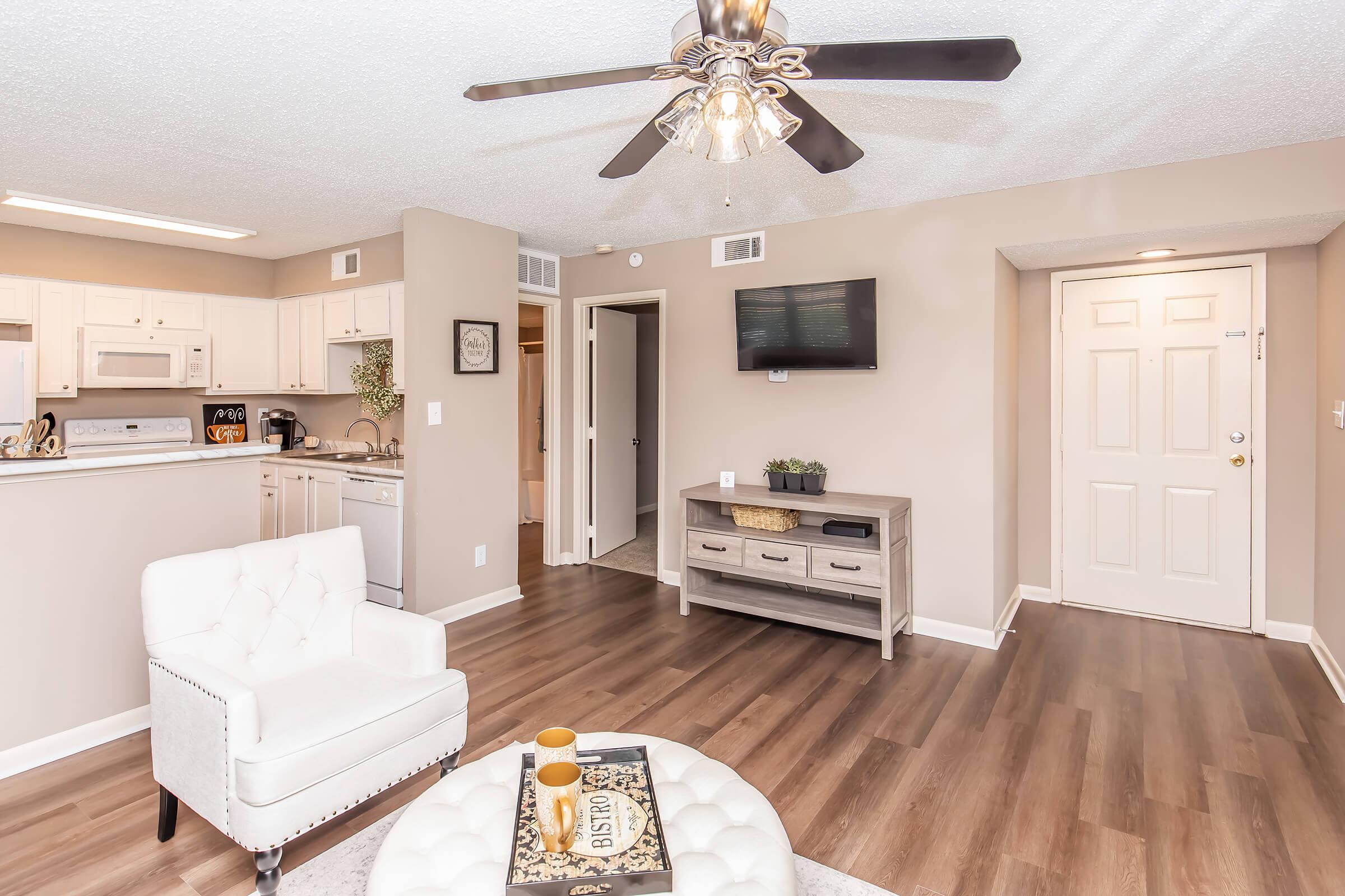 Interior view of a modern living space featuring a white armchair, light wood flooring, and neutral-colored walls. A ceiling fan hangs above, and a small table with decorative items is visible. The kitchen area is open, with white cabinetry and appliances. A TV is mounted on the wall, and a doorway leads to another room.