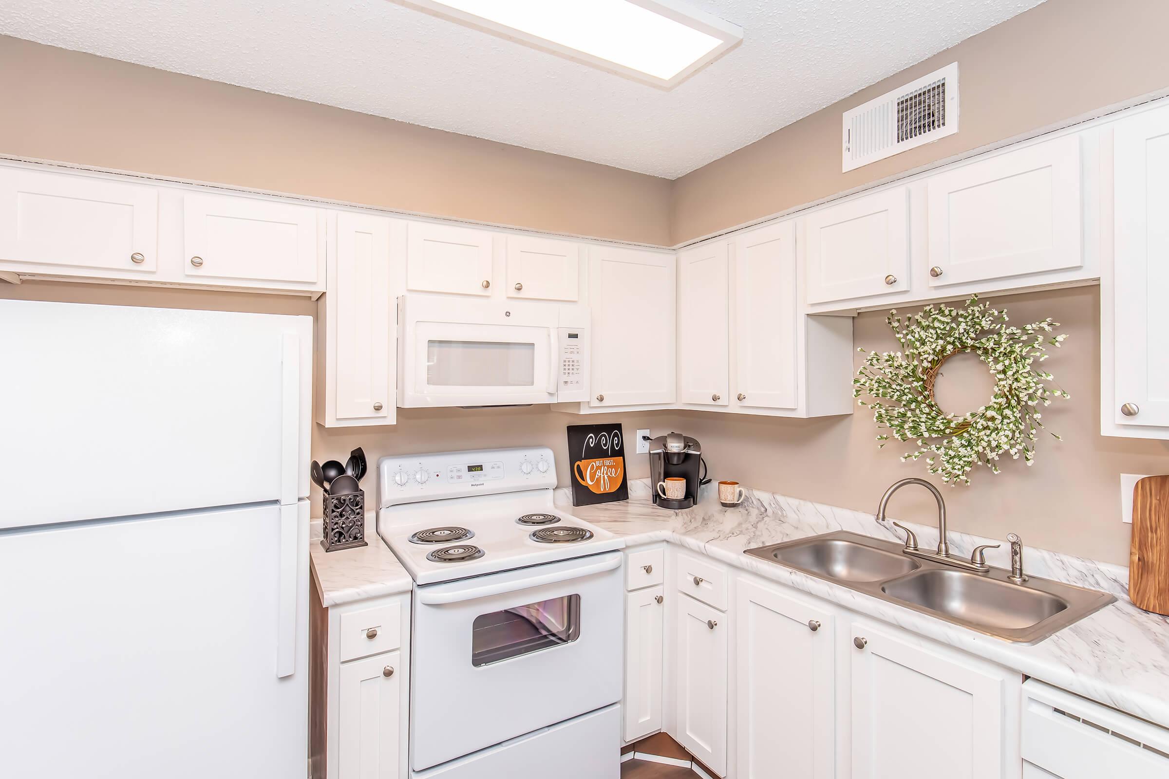 A modern kitchen featuring white cabinetry, a white refrigerator, a stove, and a microwave. There's a double sink with a marble countertop, a coffee maker, and decorative items including a green wreath on the wall. The space is well-lit and organized, creating a fresh and inviting atmosphere.